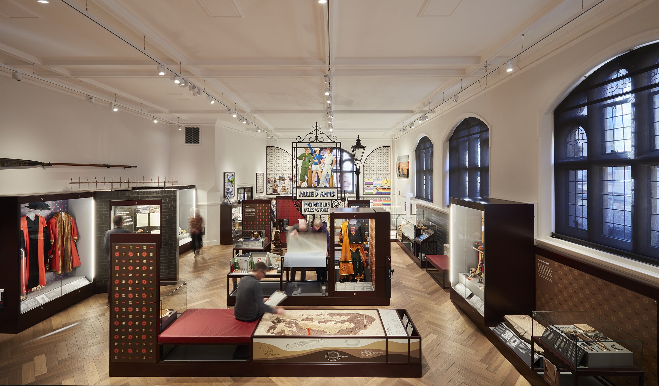 Showcases in a gallery displaying artefacts and costumes. A Victorian lamppost and historic pub sign stand taller than the other displays in the centre of the room. In the foreground a man sits by a sandbox engaging in an archaeological dig activity