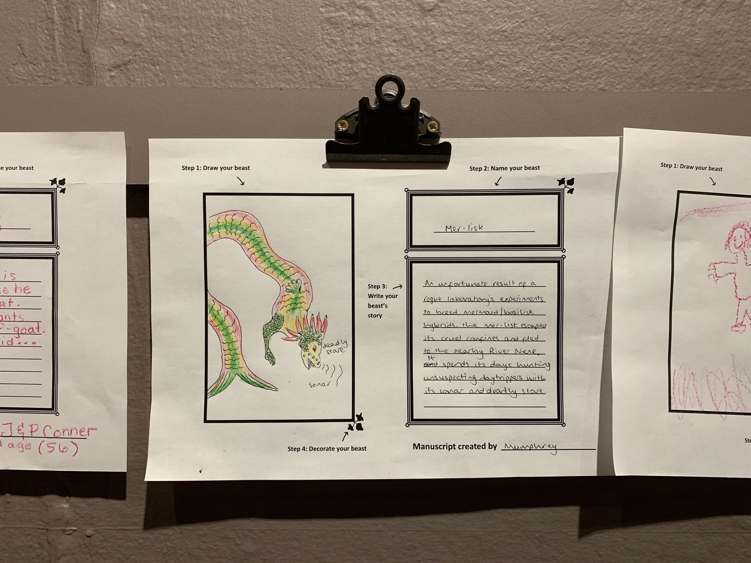 A visitor's colourful drawing of a dragon-like creature with a fish tail, which they have called a "mer-lisk," a cross between a mermaid and basilisk. The drawing pinned to a grey wall.