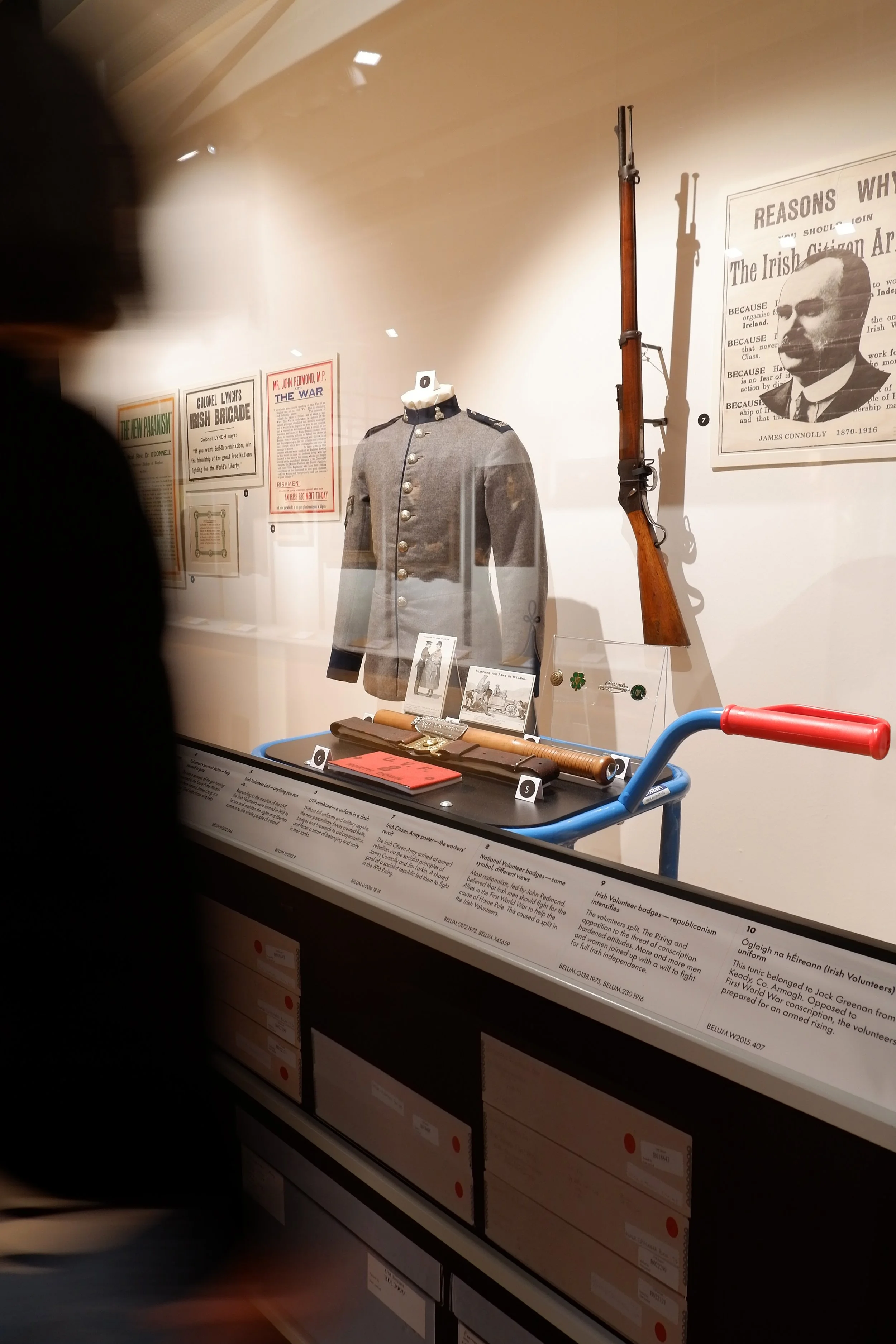 A visitor looks at a display of objects behind glass, including a grey military jacket and a selection of objects on an art handling trolley.