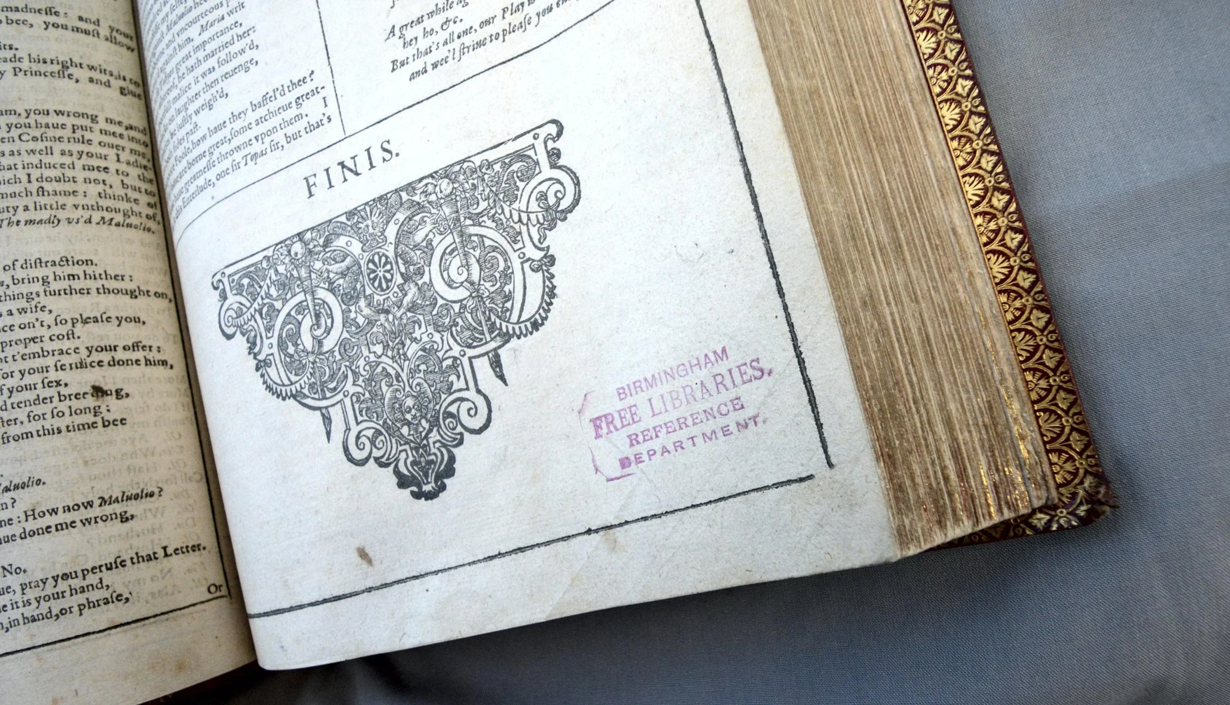 The corner of an open, ornately-bound book. The word “FINIS” is printed above a large decorative motif, signifying the end of a play. Next to it, a purple rubber-stamped text reads “Birmingham Free Libraries Reference Department”