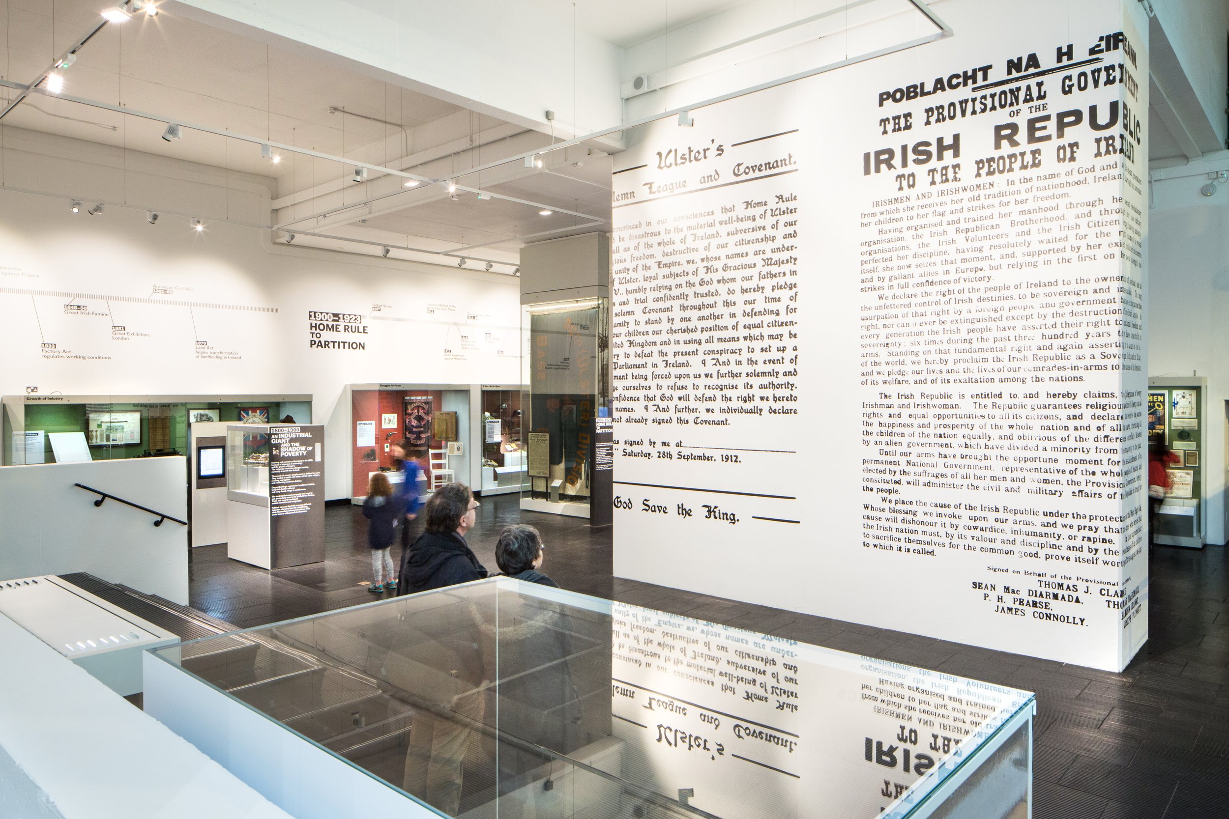 Gallery with showcases, text panels and a large timeline running around the top of the room. In the centre, two visitors look at a tall freestanding wall featuring large reproductions of the Ulster Covenant and the Proclamation of the Irish Republic.