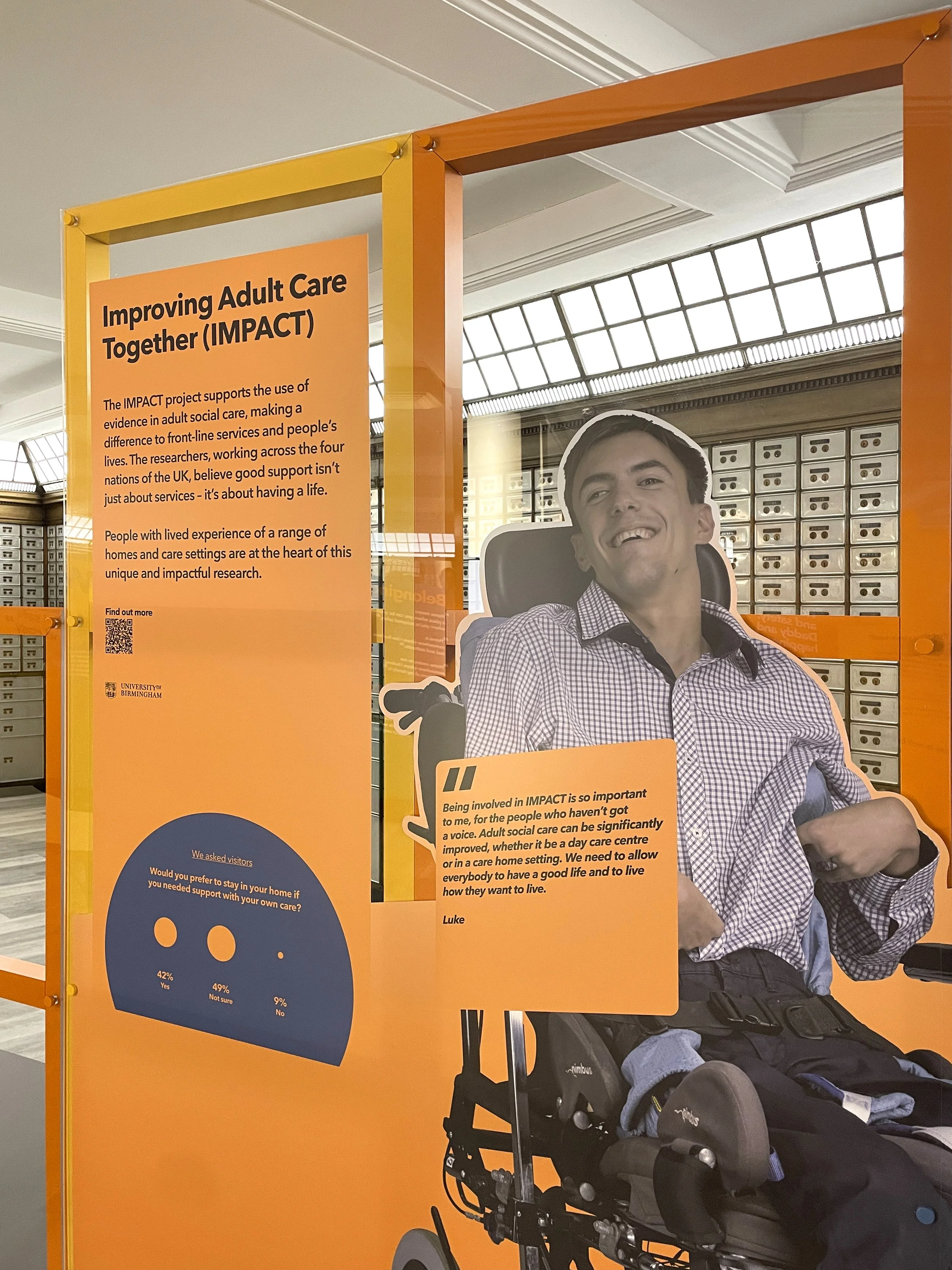 A large cut-out image of a smiling man in a wheelchair exhibited on an orange and yellow frame. There’s a quote from the man on an orange square on the image. On the left, an orange text panel is titled “Improving Adult Care Together”