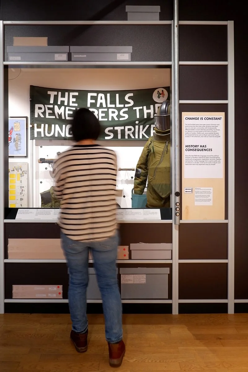 A woman looks at display of objects, including a fabric banner with the words 'The Falls Remembers the Hunger Strikers' and an army-green helmeted uniform.