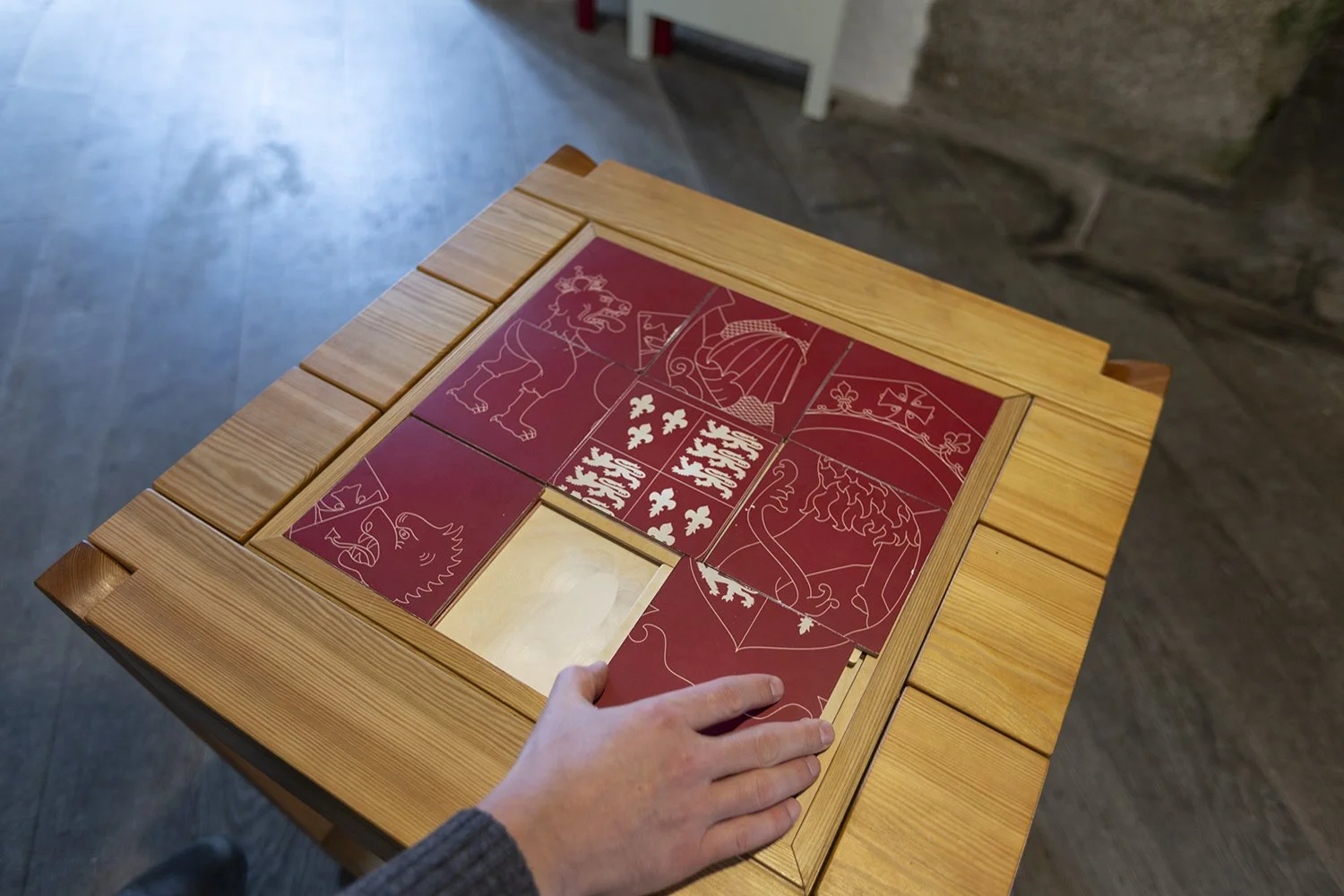 A person’s hand moves a red tile in an image puzzle which is inset into a wooden table. The red puzzle tiles feature white line illustrations and must be rearranged to form a picture.
