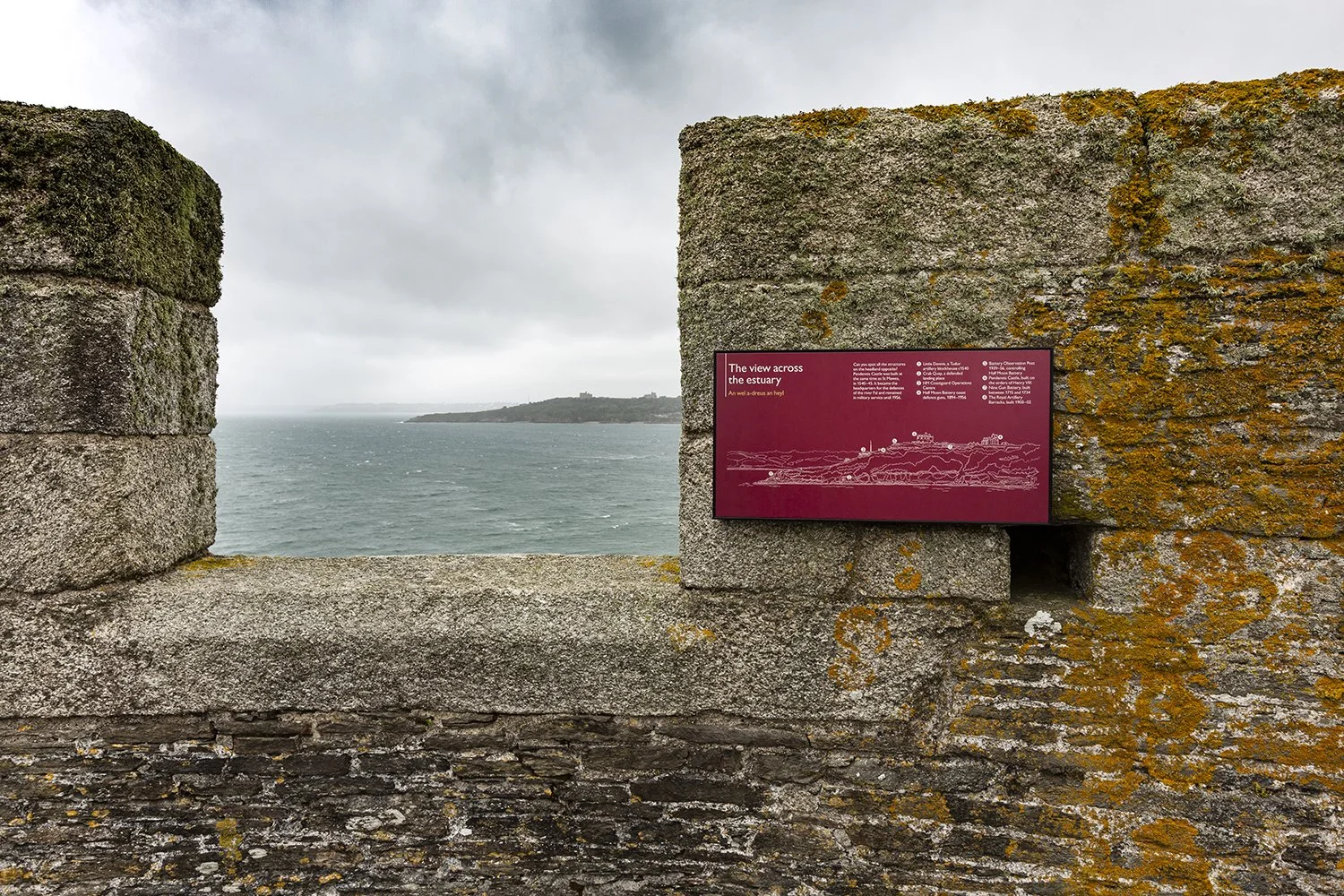 View of a headland in a grey-blue sea, seen through the square gap in a mossy stone wall. A red label is mounted to the right of the gap titled “The view across the estuary”, with a line illustration of the view and key locations indicated.