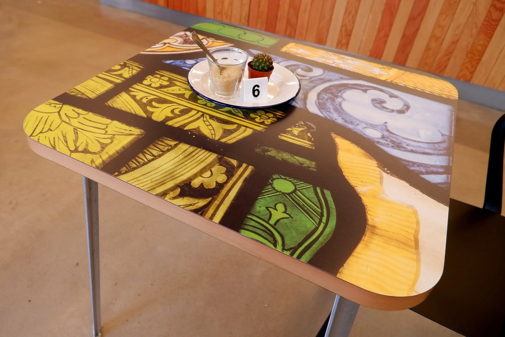 A cafe table printed with details of a colourful stained glass window. On the table is a white plate with a glass of sugar and small cactus.