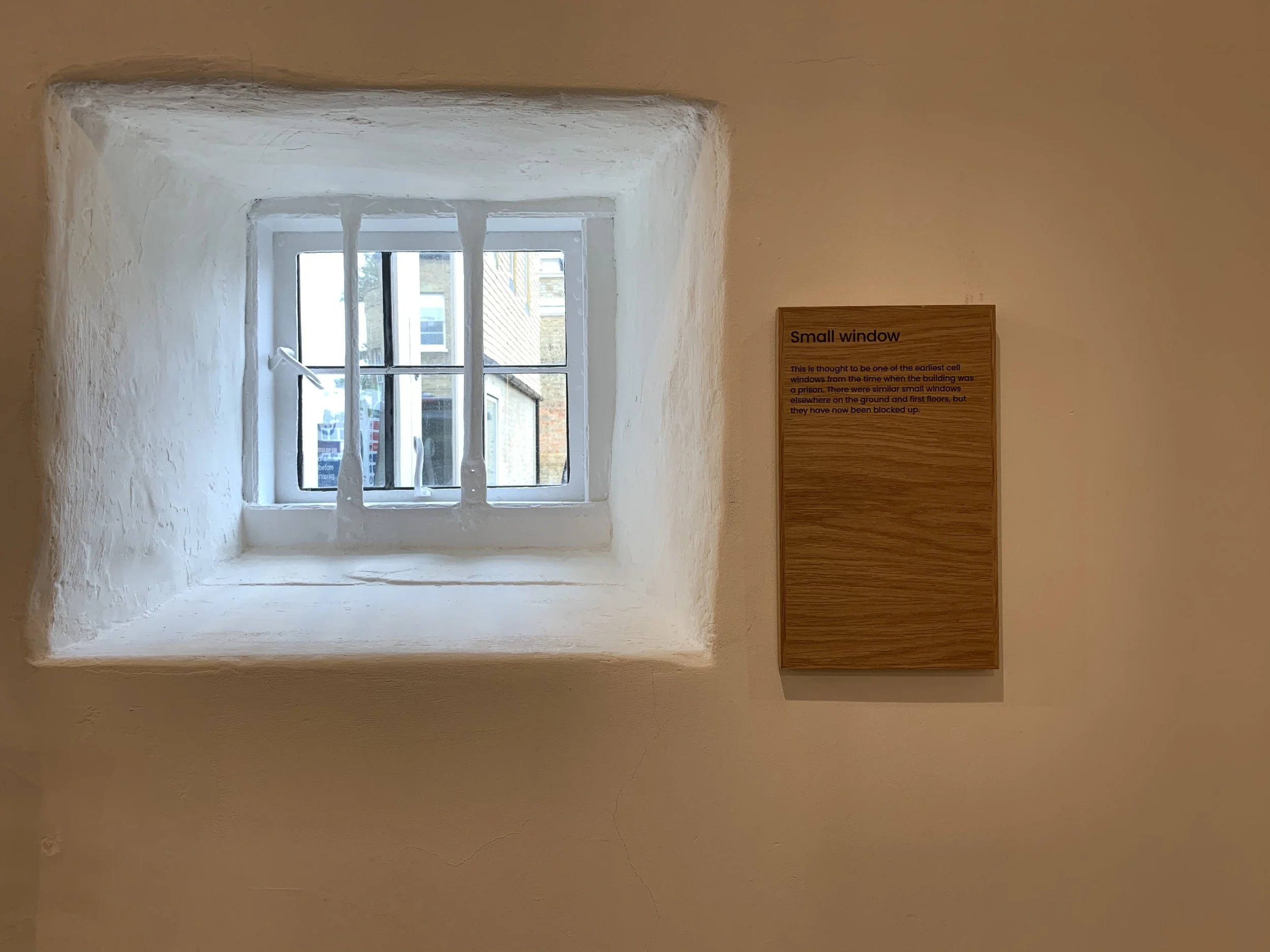 A small square window in a thick white wall. It has two metal bars on it. Next to it is a wooden text panel titled “Small window” that explains this was an original cell window in a prison.