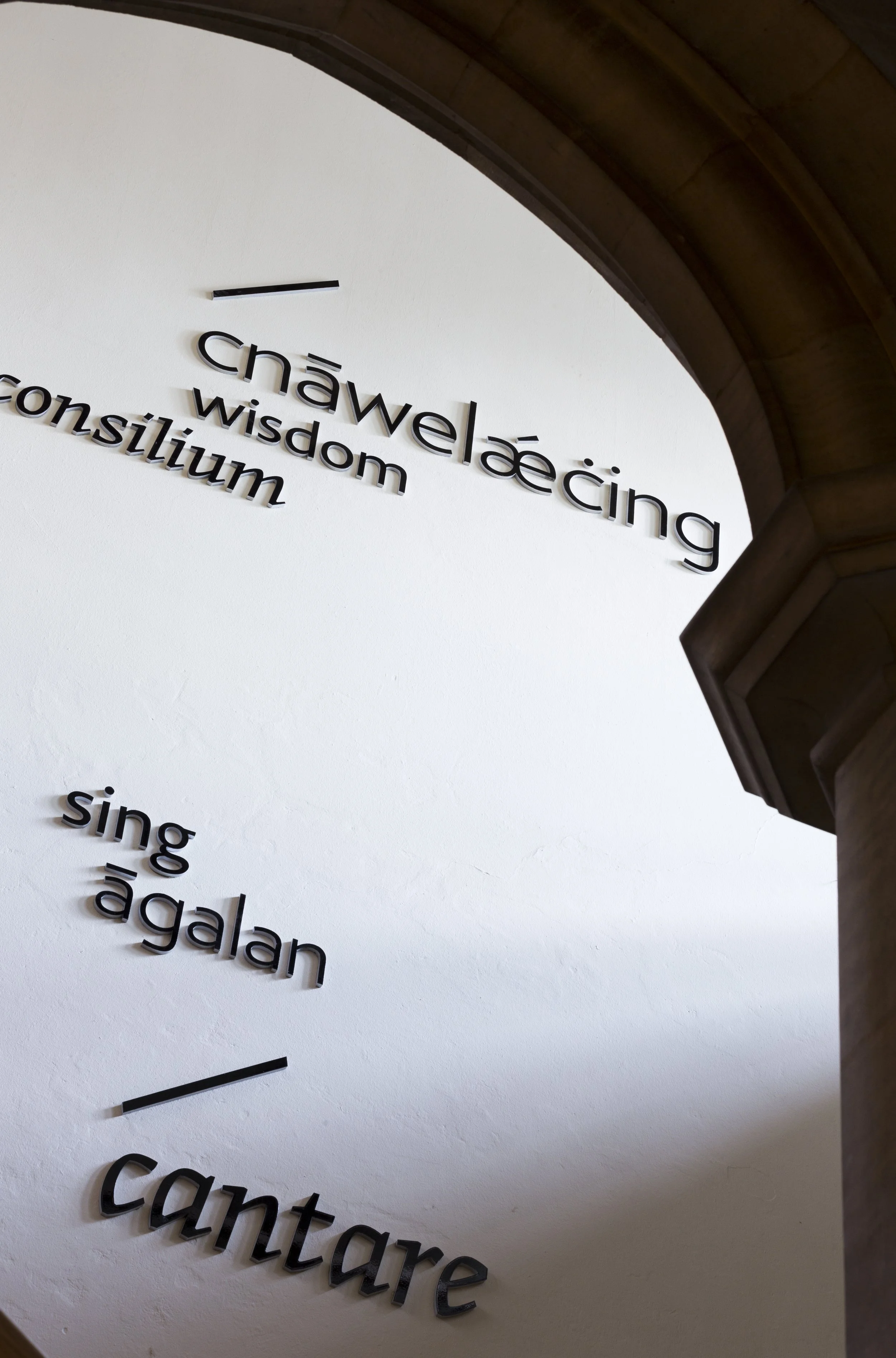 Looking up at a white wall from beneath an archway. Large words in different languages, fonts and sizes are mounted on the wall as aluminium lettering, casting gentle shadows. They include translations for the words "sing" and "wisdom."
