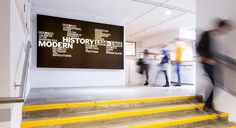 Blurred figures walk past the title panel outside the exhibition entrance, at the top of a concrete staircase. The graphics are printed in white text on a large black panel, with the title “Modern History 1500-1968” surrounded by key dates.