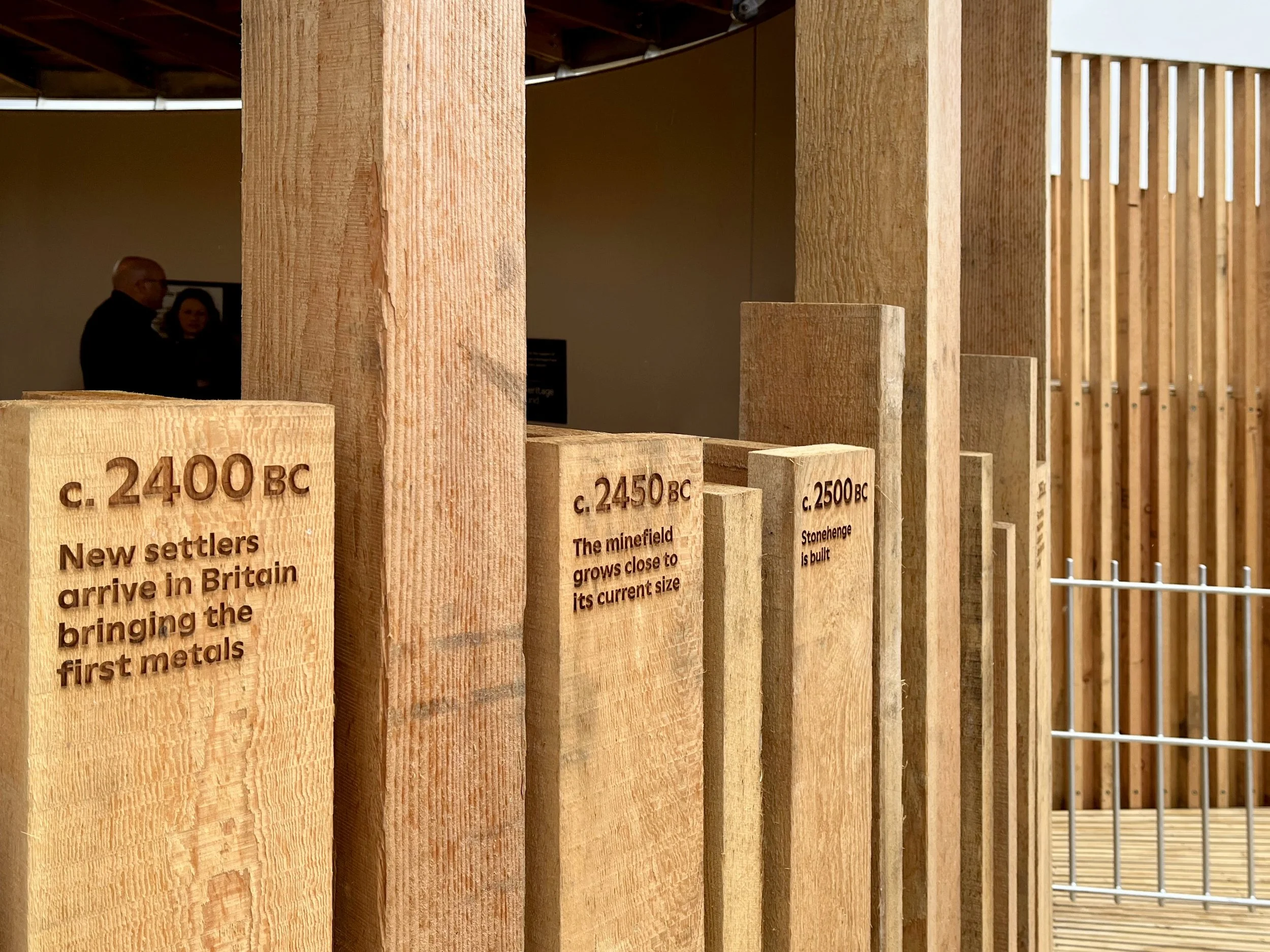 At Grime's Graves in Norfolk, a wooden structure is made off timber posts which have engraved dates and interpretation. At the background of the image there are people mingling in front of a graphic panel.