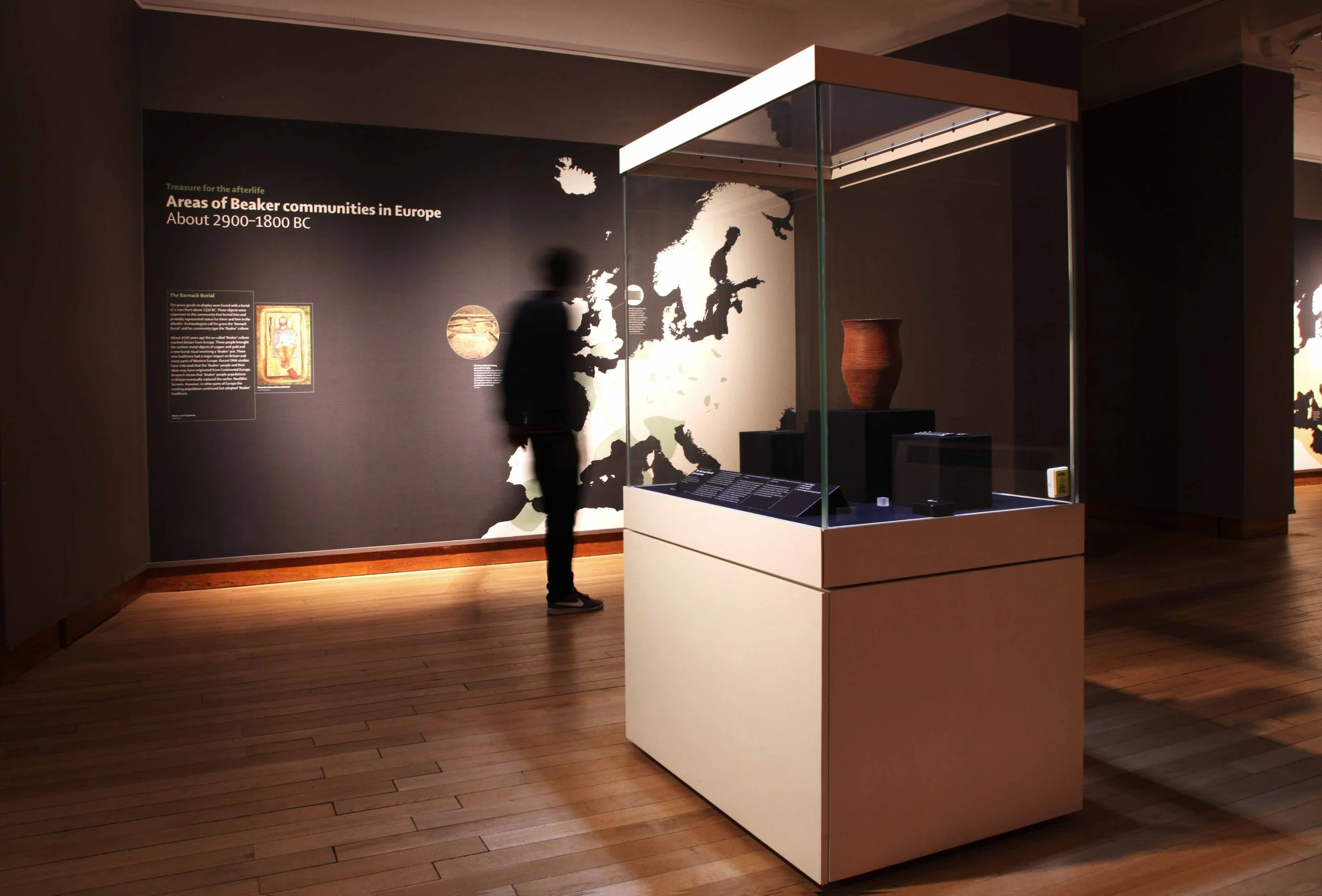 A visitor walks through a gallery past a wall-sized map of Europe showing locations of Beaker communities between 2900-1800 BC. In the foreground a white showcase displays a clay vase.