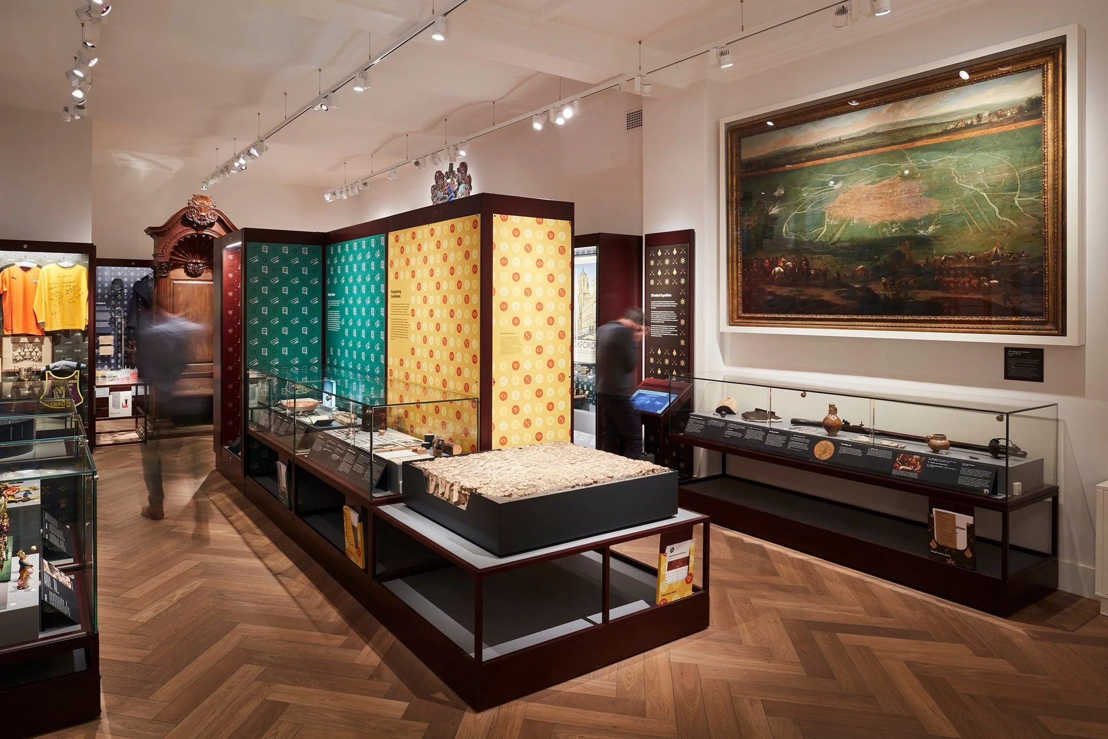 Museum objects in showcases with colourfully patterned back panels. Visible objects include a model carousel and football shirts. Two helmeted figurines holding axes are mounted to the left wall above the displays.