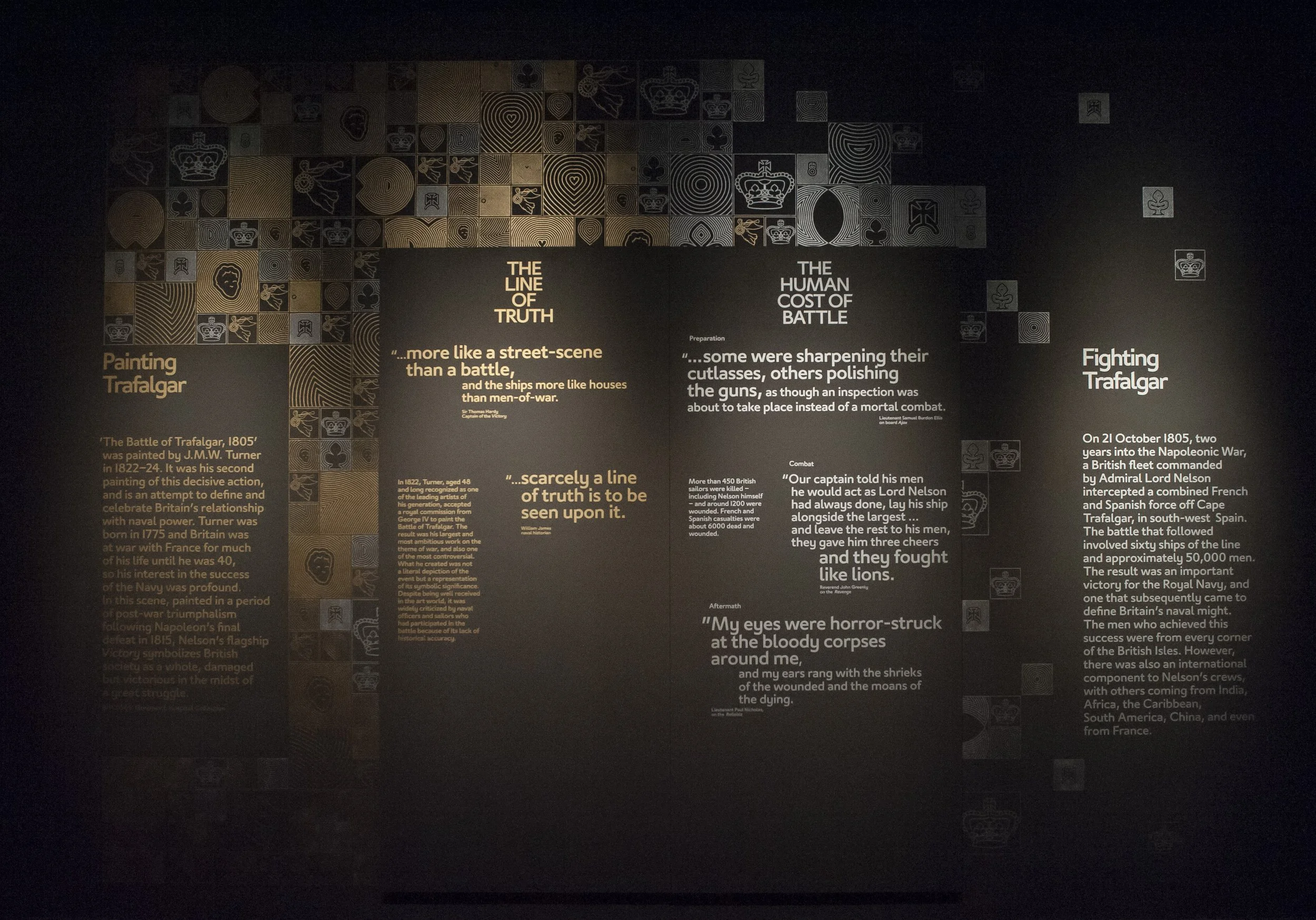 A black wall printed with gold and silver motifs. In the centre, two text panels are titled “the Line of Truth” in gold and “The Human Cost of Battle” in silver, with various quotes beneath them.