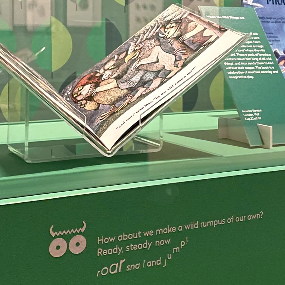 A blackboard with white text titled “At School.” A4 drawings are strung across the board, with a school desk and four chairs beneath it, and a wall-mounted showcase containing books on the left.