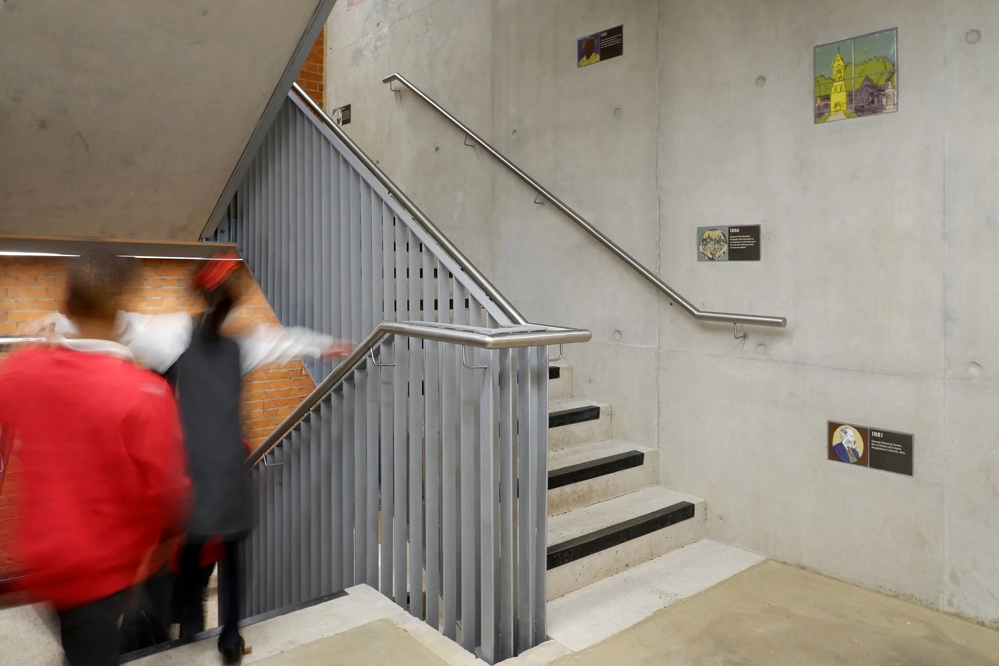 Two children walk down a staircase. Behind them, ceramic tiles are inset into the cast concrete wall, featuring colourful illustrations and text.