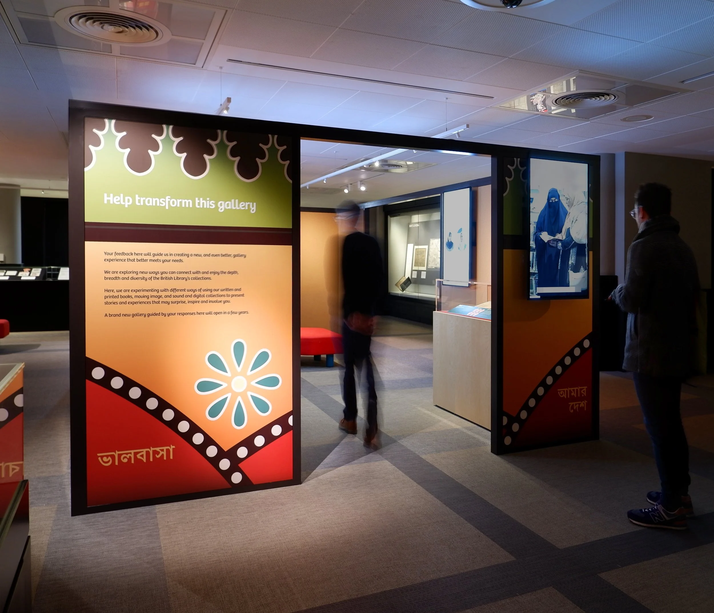 A colourful rectangular structure stands in the middle of a gallery at the British Library. Its graphic surface has shapes of flowers and other symbols related to Bangladeshi culture. A man is watching the introductory video.