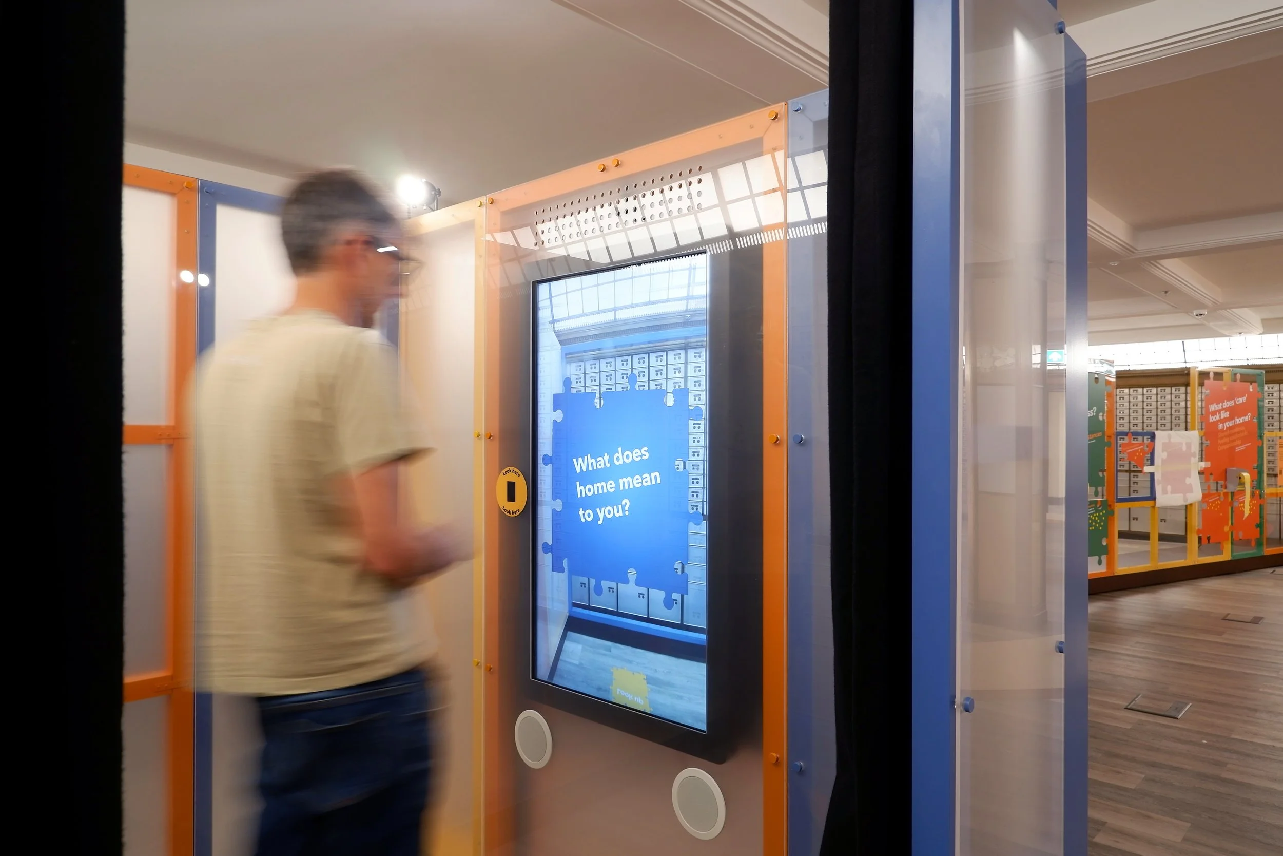 A man stands in a booth facing a video screen with a blue jigsaw piece on it, and the question “What does home mean to you?”