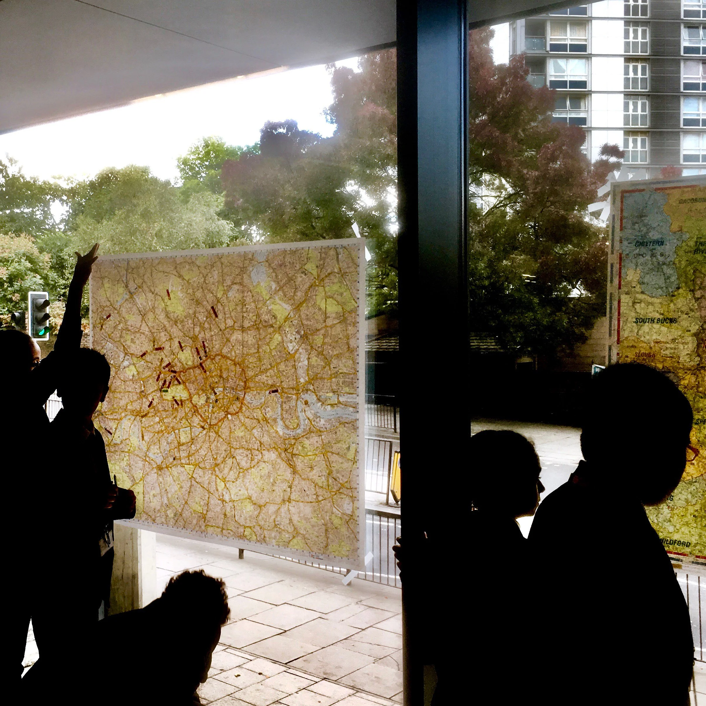 Children stand in front of large maps which are pinned to a window. The maps have markers on indicating different locations.