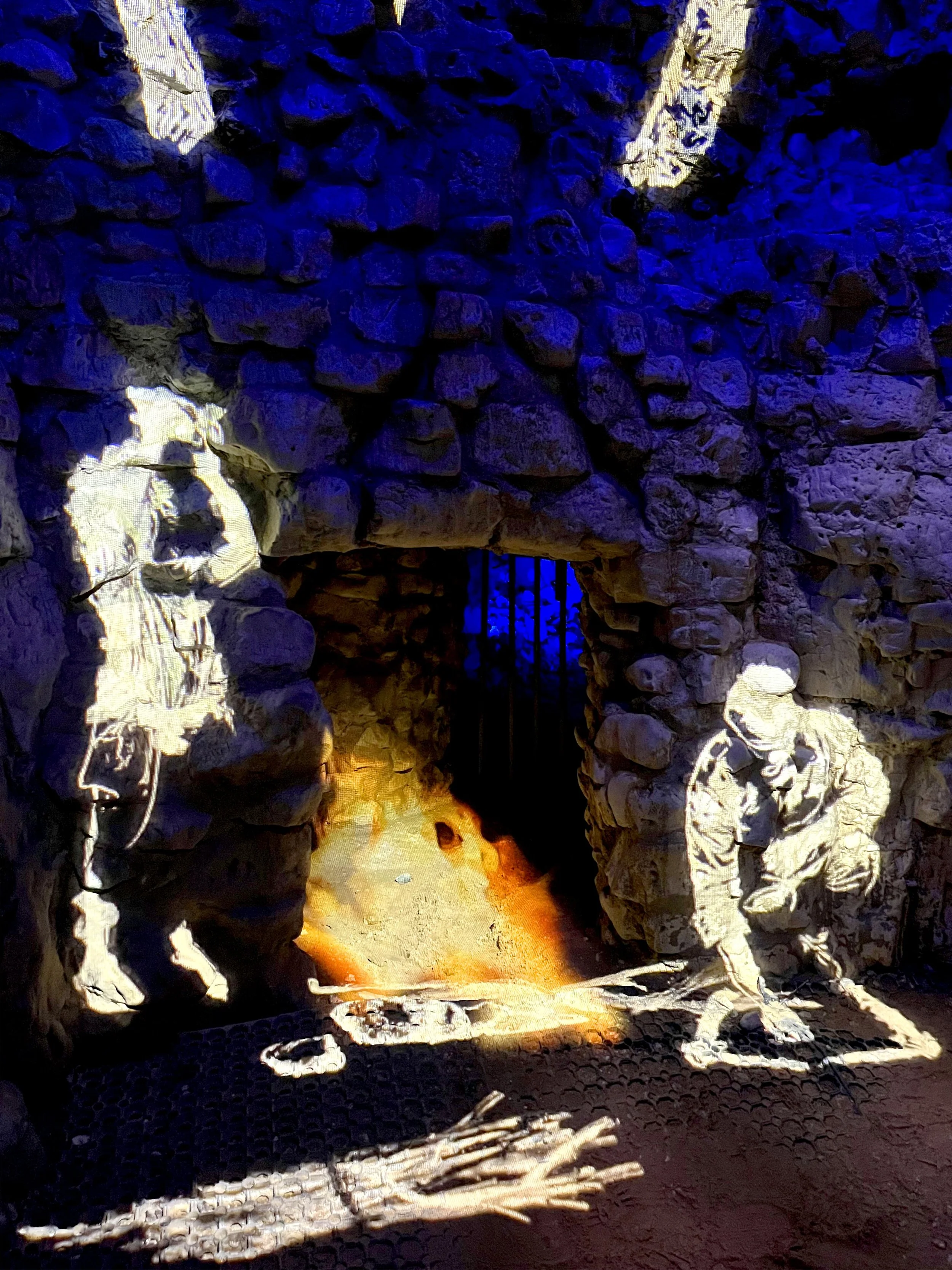 The photo shows the interior of one of a vast complex of prehistoric flint mines at Grime's Graves in Norfolk. Projections on the stone wall showing a fire pit and two human figures; one standing and one sitting on the floor.