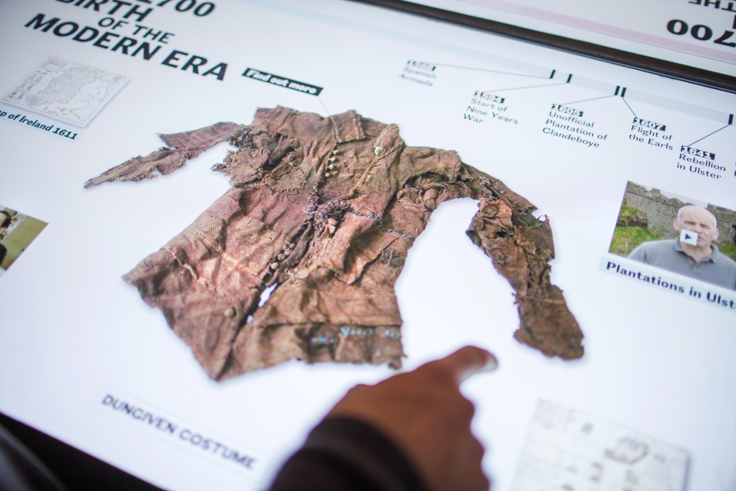 A hand touches an interactive digital display. On screen is an image of a brown tattered shirt titled “Dungiven Costume.” Onscreen options include a timeline of events from 1500-1600s, a 1611 map of Ireland and a video titled “Plantations in Ulster.”
