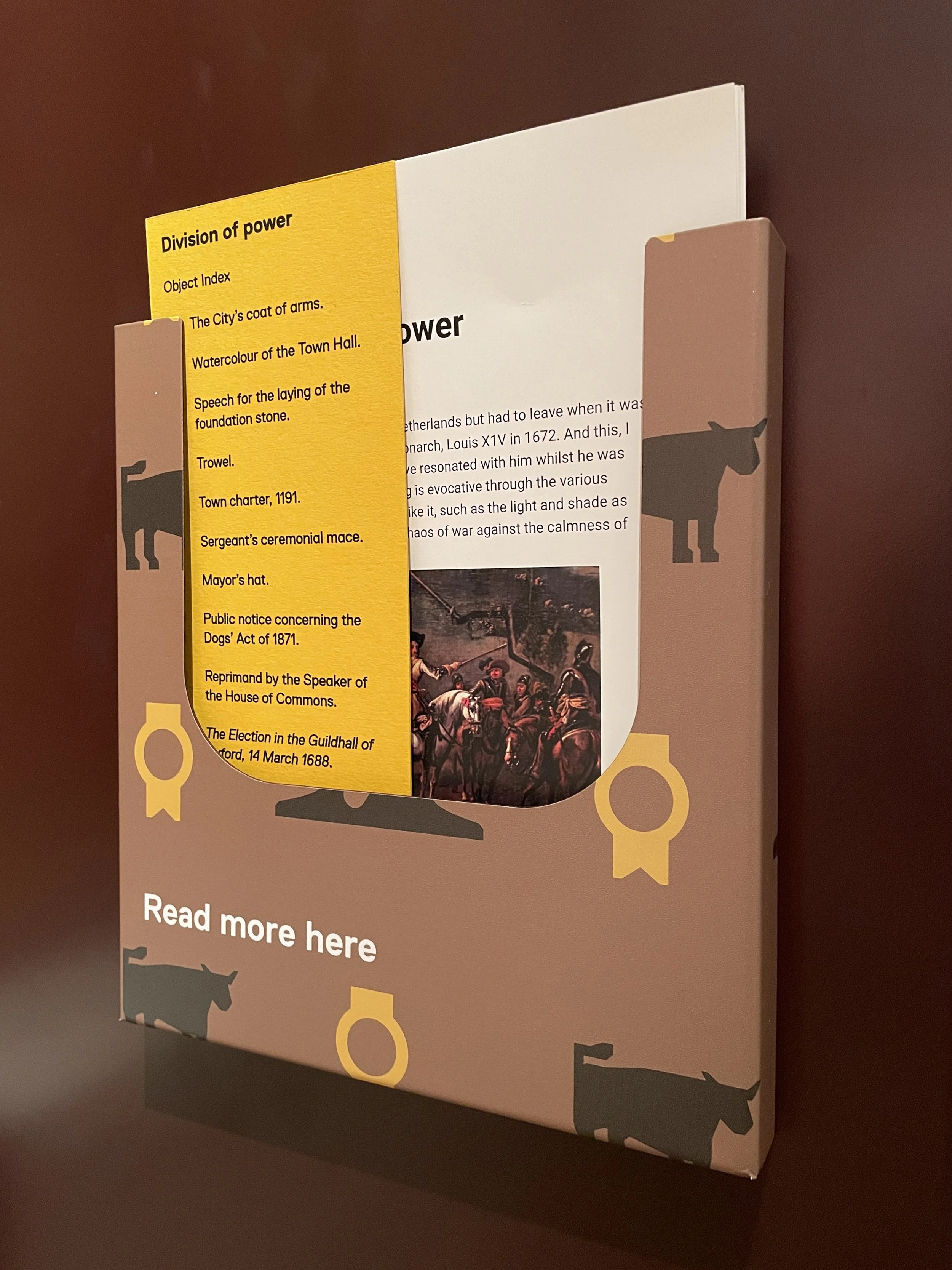 A folded metal holder containing white and yellow papers with printed text and images. The holder is decorated with silhouettes of cows and includes the caption 'Read more here'.
