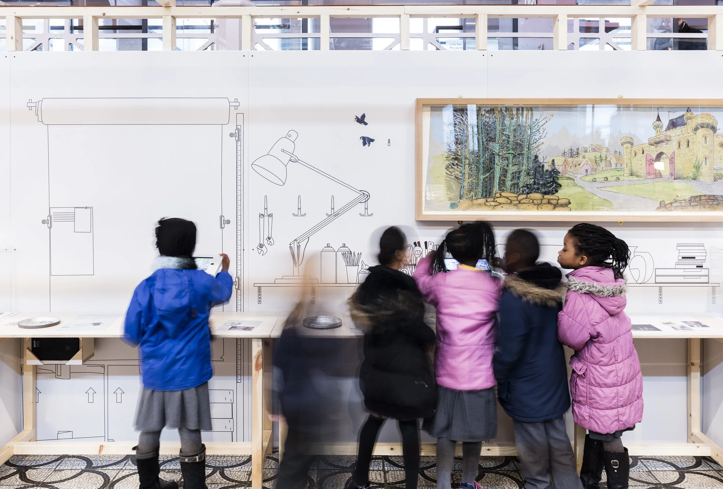 Six schoolchildren with purple & blue coats play with iPads on a table with their backs to us. Behind them a white wall is decorated with line drawings of a lamp, spraypaints, pencils and piles of books, and a framed painting of a forest and castle.