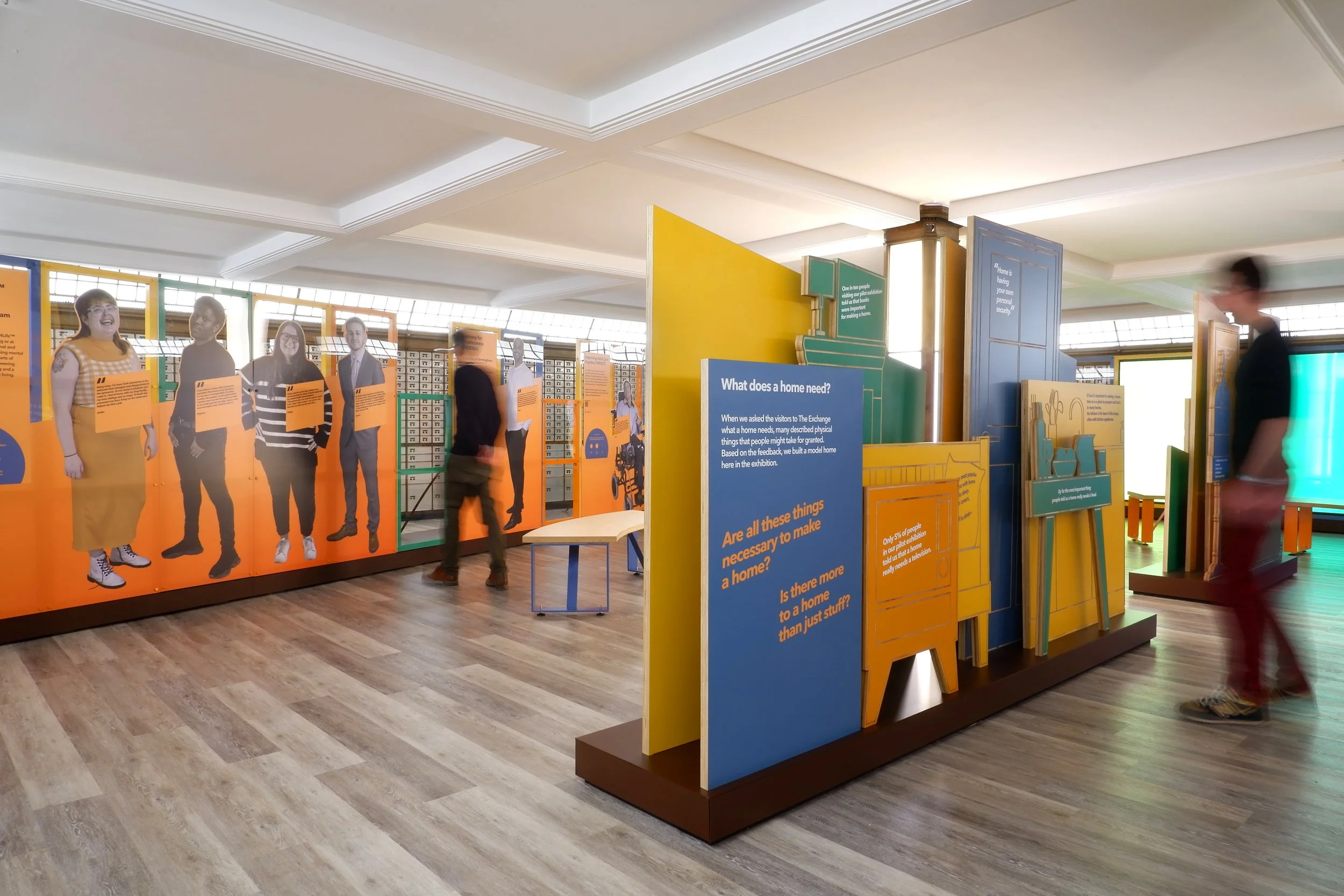 Two visitors look at exhibits in a gallery. Lifesize images of smiling people are printed on orange panels with quotes. In the foreground a freestanding display of blue, orange, yellow and green shapes ask questions about what a home needs.