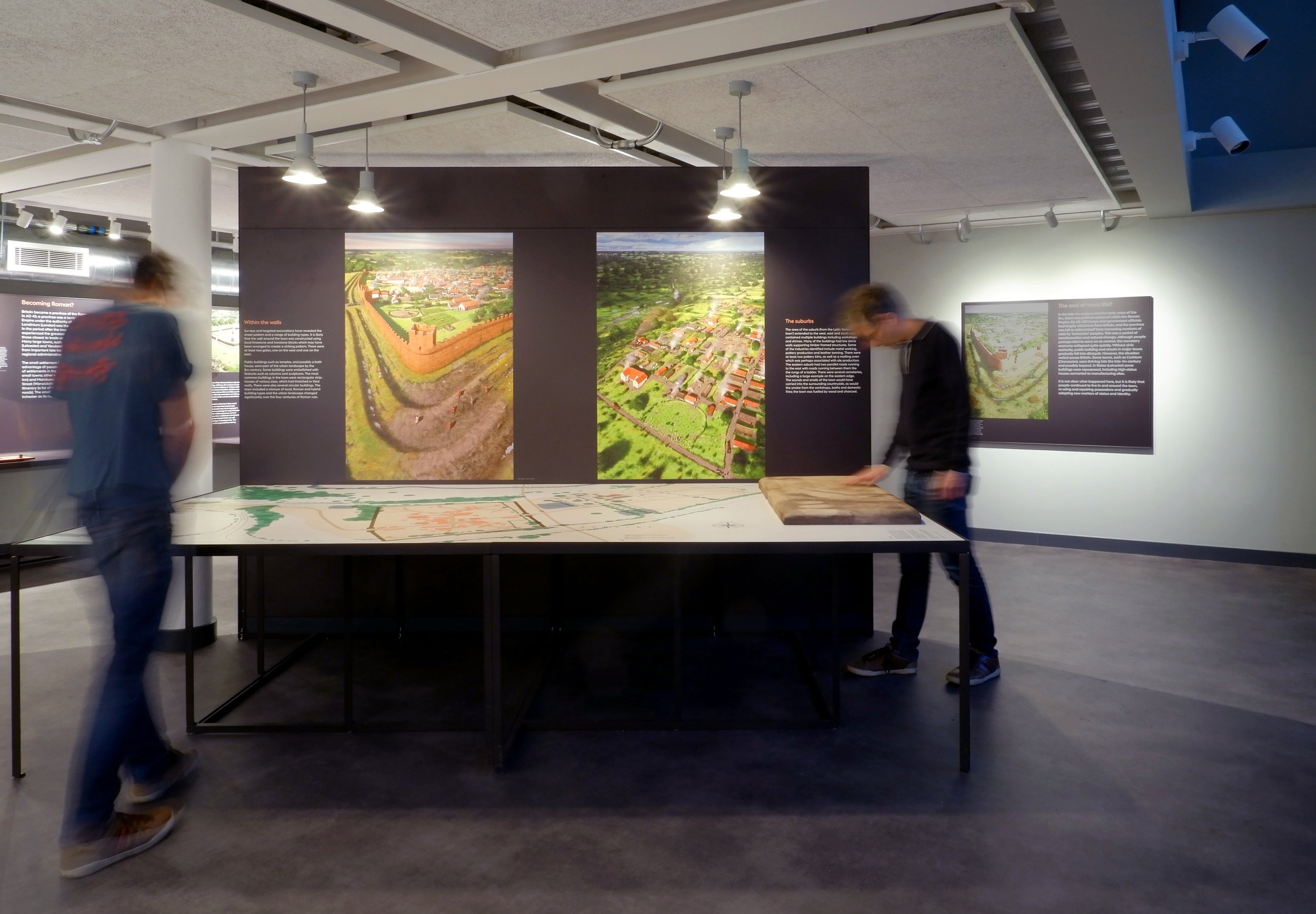 Two men look down at a tabletop map of a local area. Behind the table is a black wall with two archeological illustrations showing a wall town and a suburb with white explanatory text.