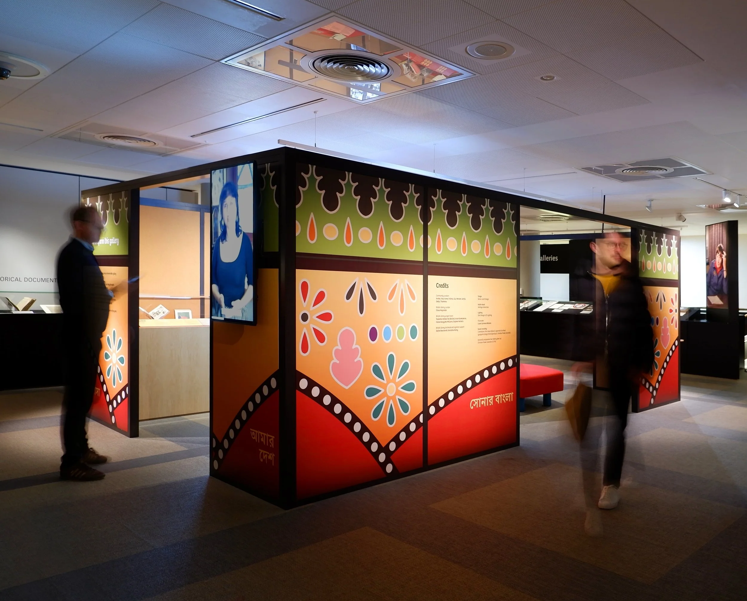 A colourful rectangular structure stands in the middle of a gallery at the British Library. The structure's graphic surface is colourful and bears shapes of flowers and other symbols related to Bangladeshi culture.