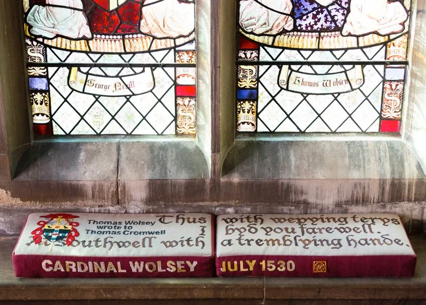 Two embroidered cushions on a stone platform beneath two stained glass windows, featuring a quote from Cardinal Wolsey to Thomas Cromwell dated July 1530. The white and red colour scheme and gothic text style of the cushions echo the windows.