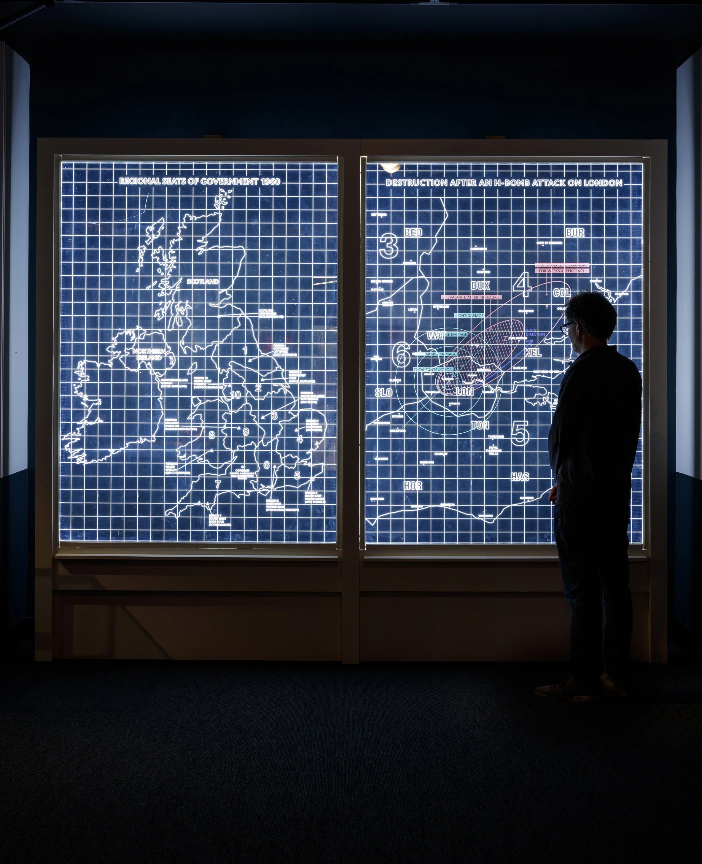 A visitor stands in a dark space looking at two large back-lit line-drawn maps of the British Isles against a gridded blue background. One map is titled “Destruction after an H-bomb attack on London,” the other “Regional Seats of Government 1960”.