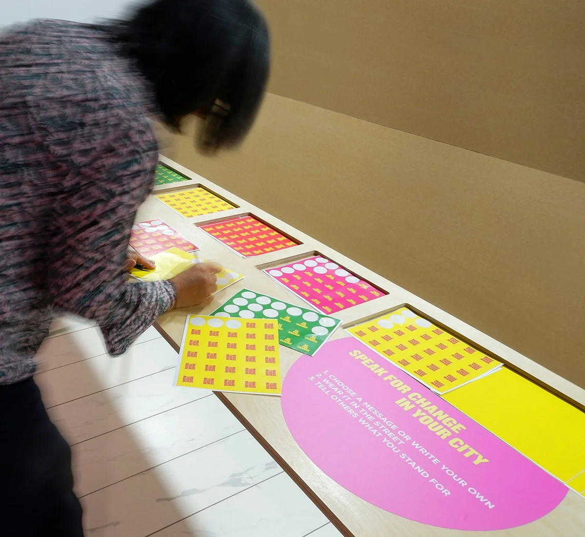 A woman takes a colourful sticker from a table with a sign reading "Speak For Change In Your City"