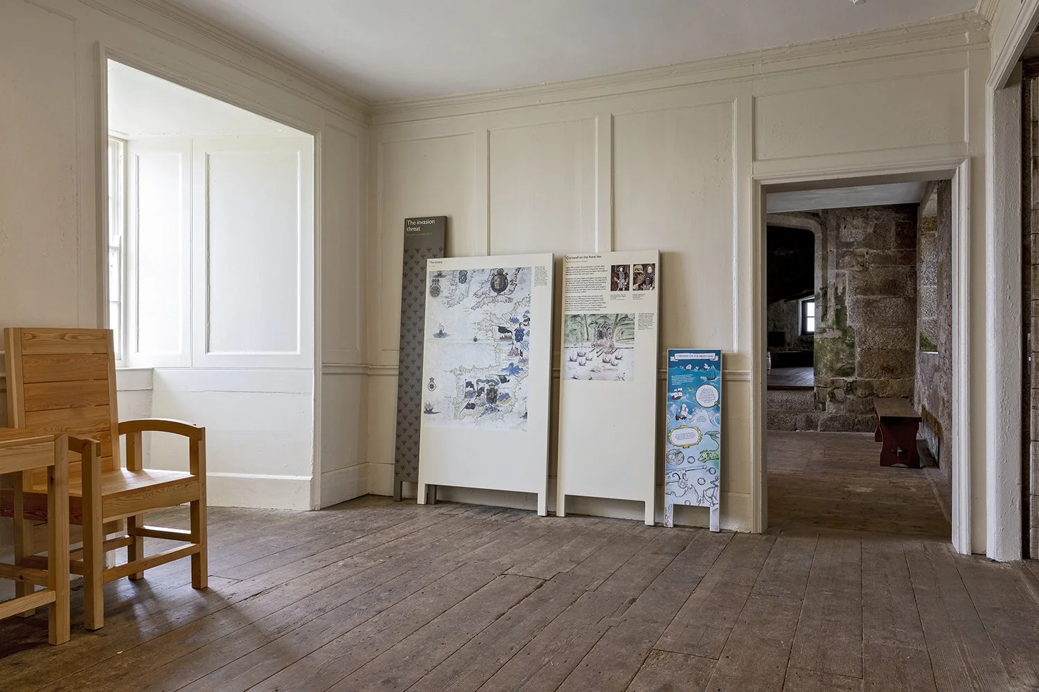 Exhibition panels leaning against a white wall in a room with wooden floorboards. The panel is titled “The invasion threat” with maps, images and text. A recessed window on the left gives natural light. A doorway leads to further rooms ahead.