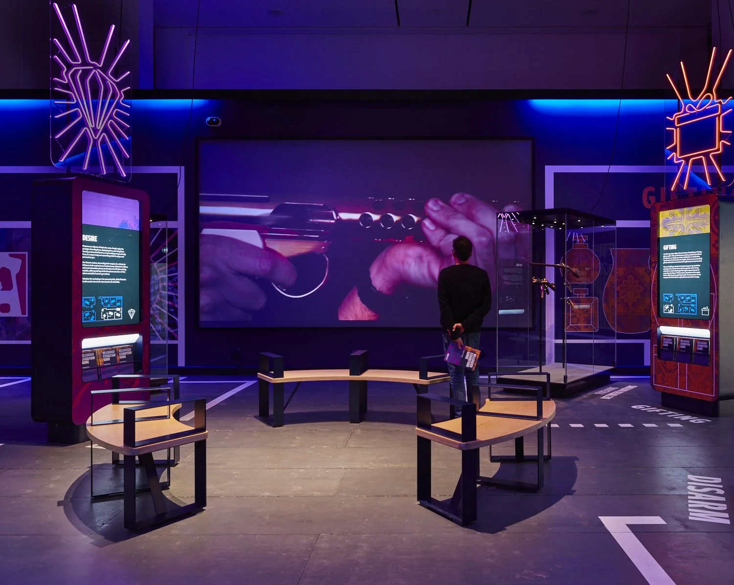 A Person stands in front of a large film projection showing a gold gun fired in slow motion. They hold an exhibition guide. In the centre are three circular benches. Two graphic introductory panels with neon lights above in the shape of a diamond.