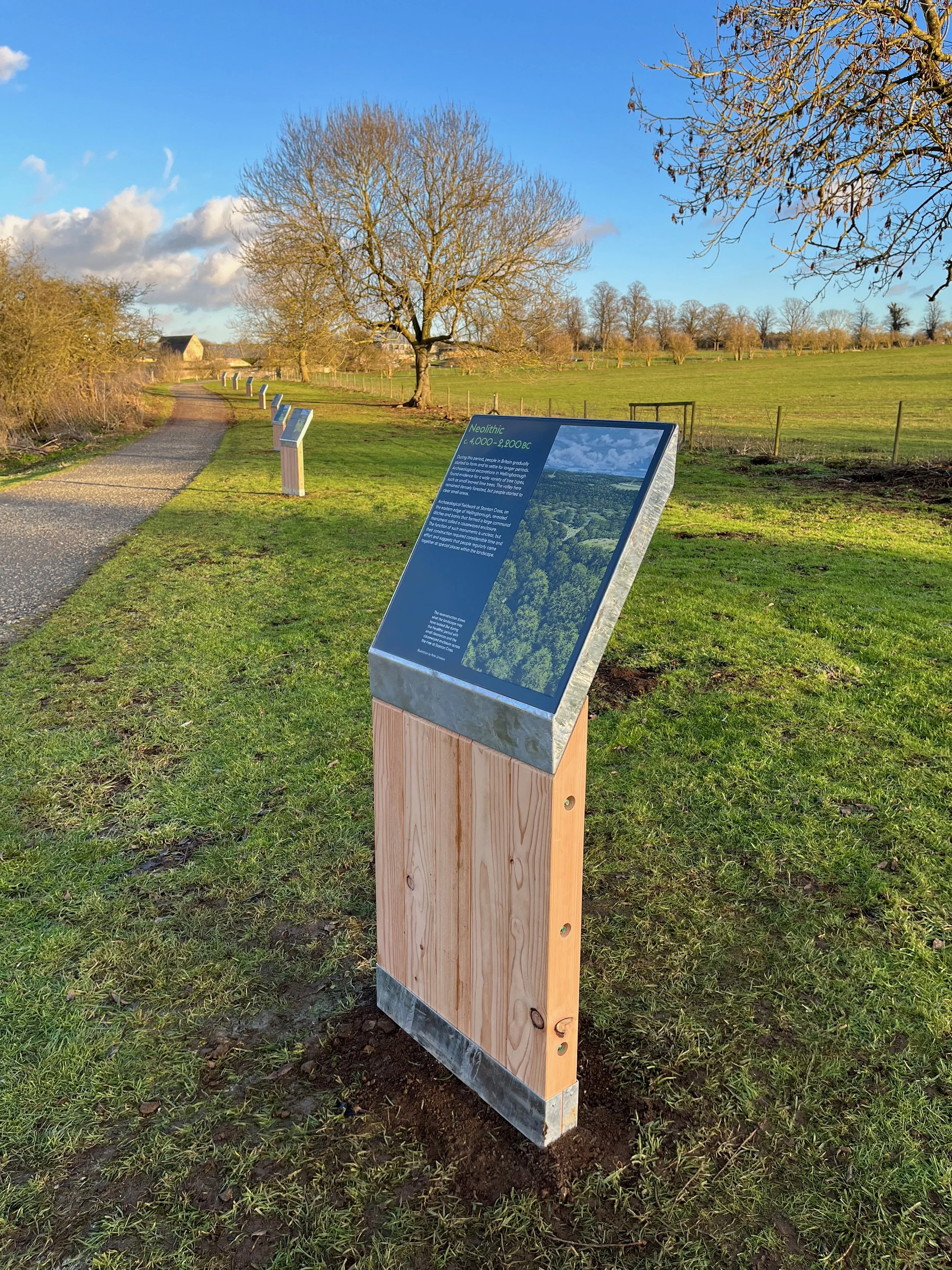 A path through a grassy landscape under a blue sky. Slanted wooden text panels are positioned at equal distances along the path. The nearest one features an aerial photo of trees with the title “Neolithic c.4,000-2,200BC."