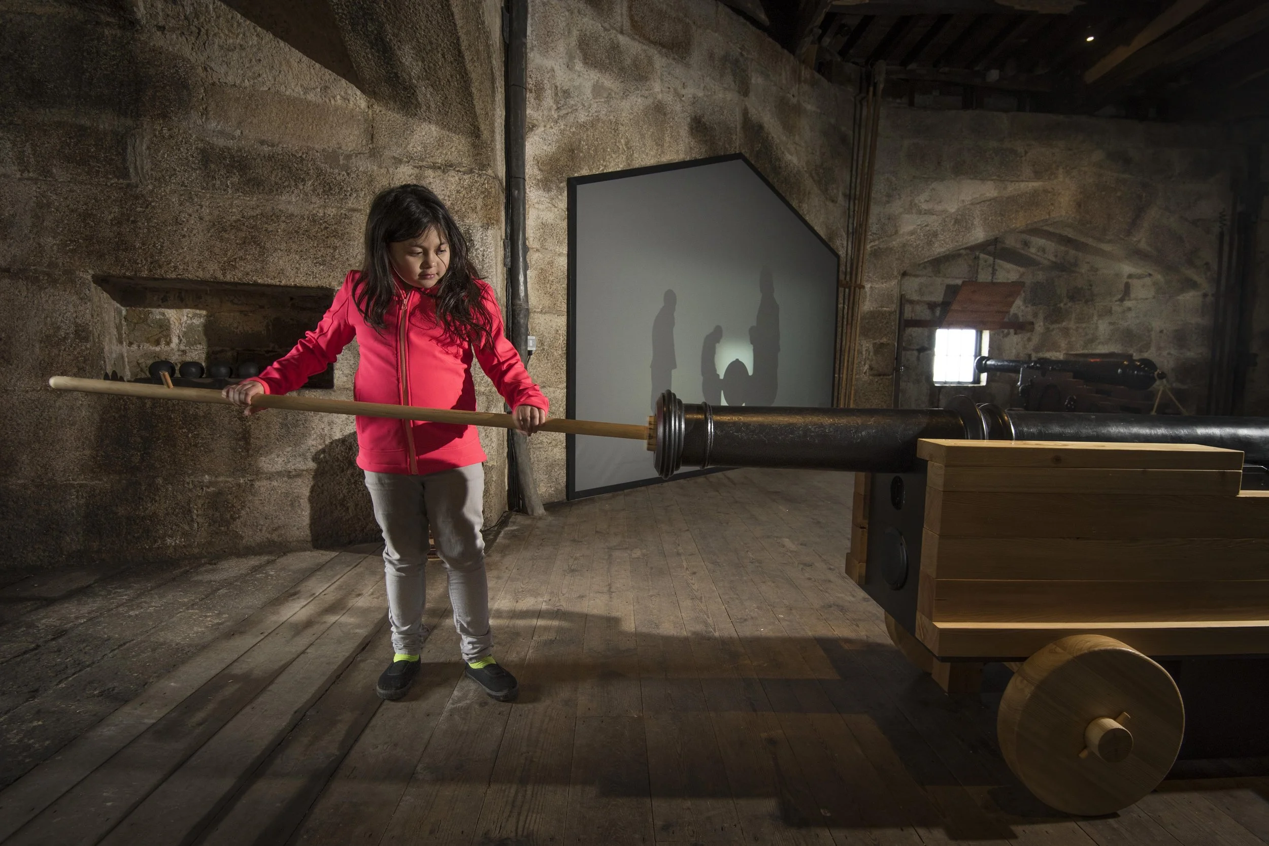 A young girl wearing a red jacket uses a pole to load a replica cannon in a room with wood floors and stone walls. Behind her is a video projection of silhouettes of men loading a cannon.