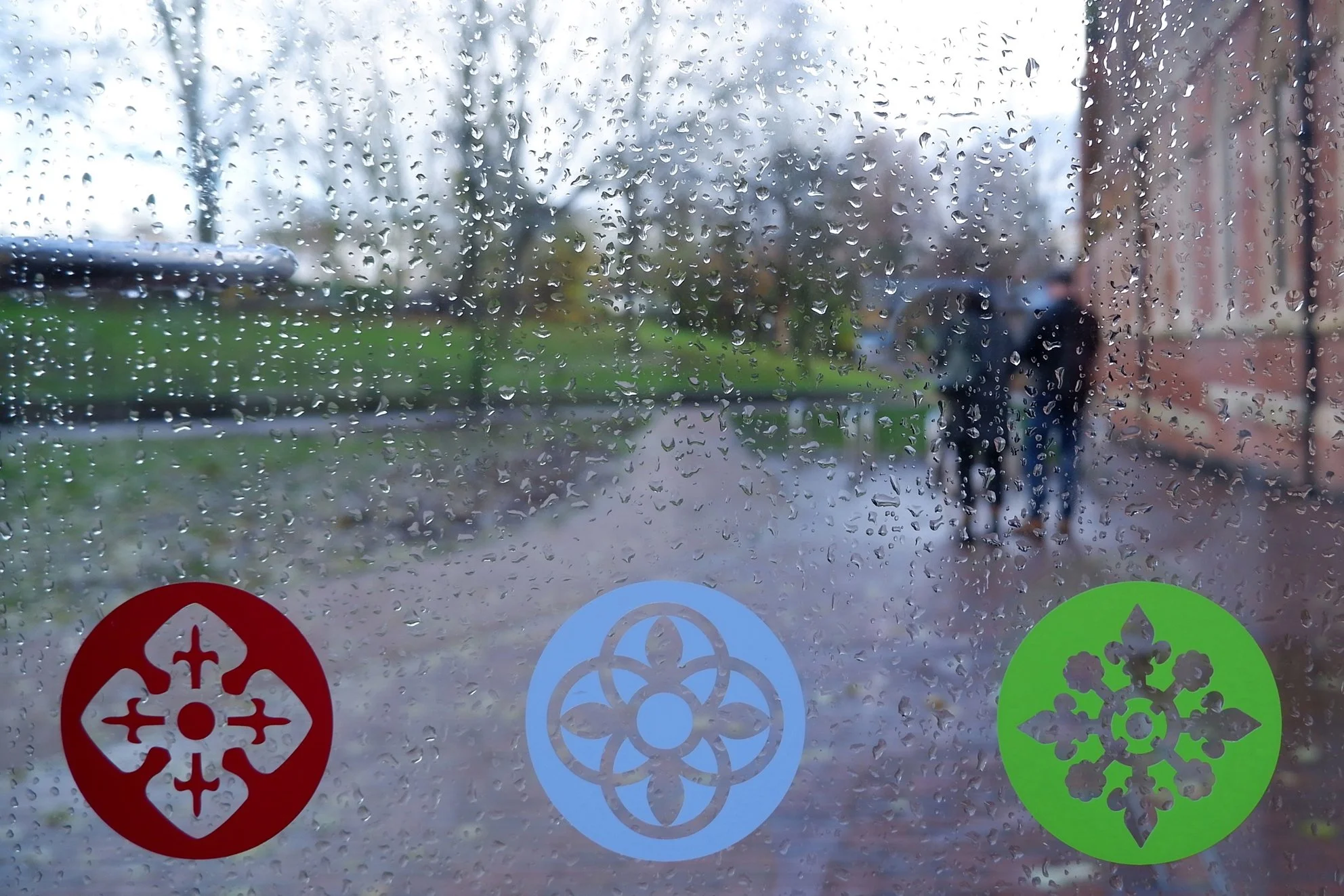 Raindrops on a windowpane with three circular stickers – red, blue and green – at the bottom of the pane, each with a different motif. The view outside is out of focus.