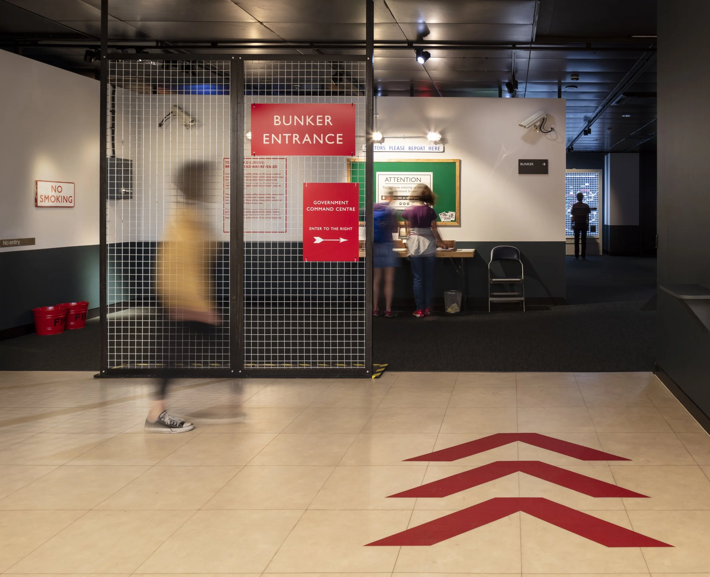 A child walks past wire-fence doors with a red sign reading “Bunker Entrance.” Red arrows on a white floor indicate the entrance to the exhibition. In the background visitors look at objects on a table.