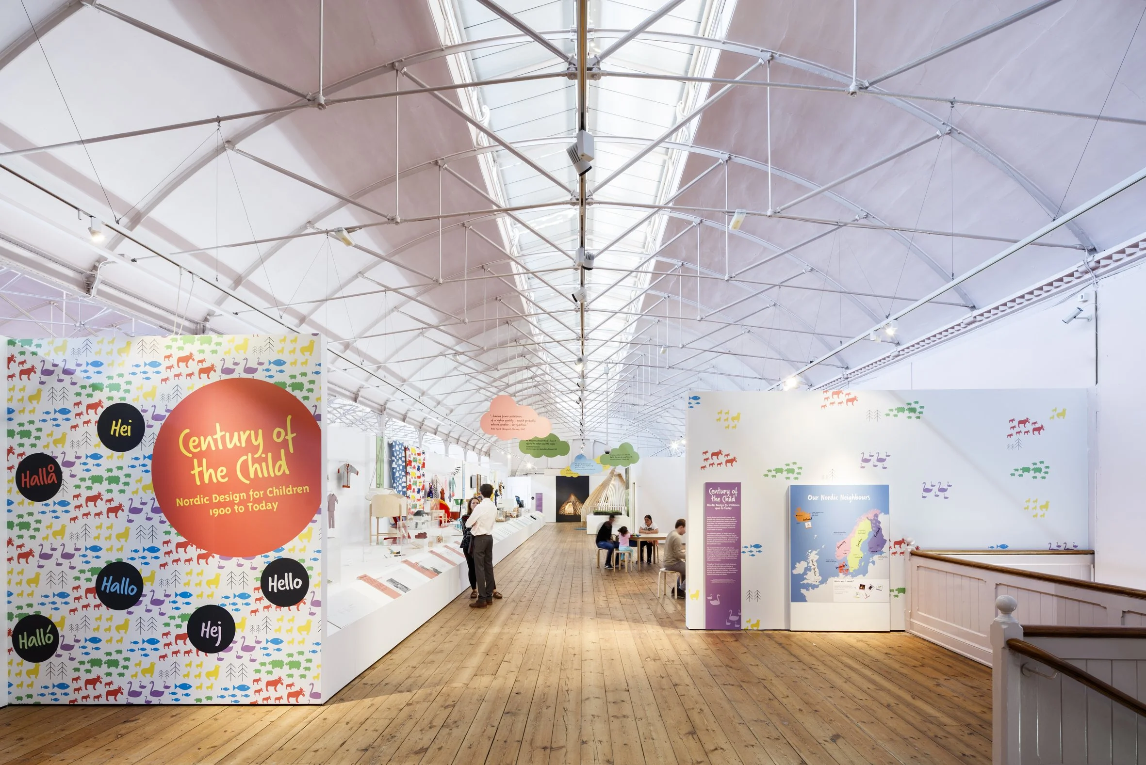 A gallery with curved ceiling and wooden floor. A long white plinth with exhibits runs down the left side. On the right visitors sit at low tables. A colourful entrance panel reads “Century of the Child” with words for hello in Nordic languages.