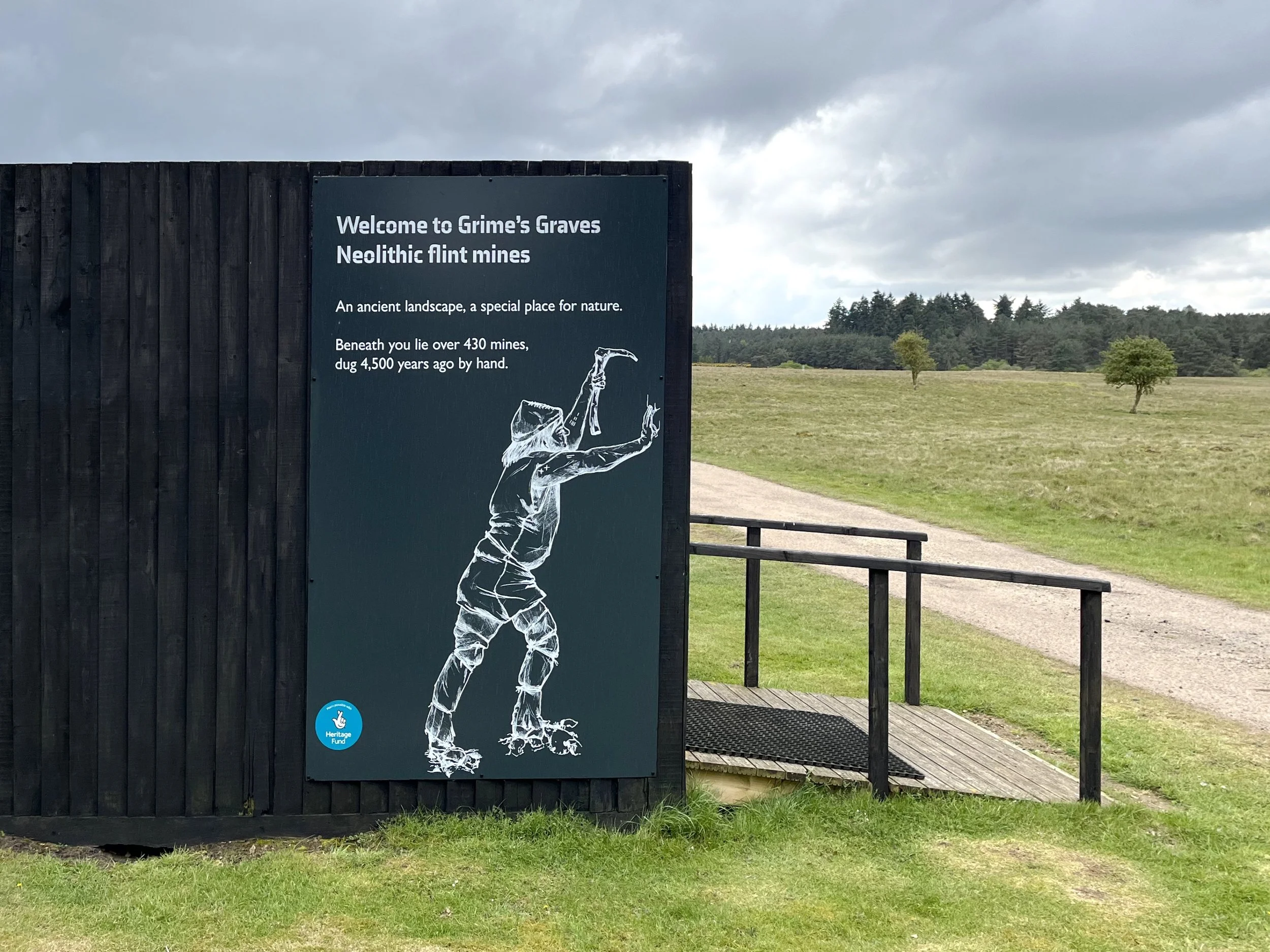 Welcome sign at the park entrance, inviting visitors with open arms to enjoy the serene beauty and recreational activities. On the exterior wall of the pavilion there is a graphic panel with an illustration of a Neolithic man holding a tool.