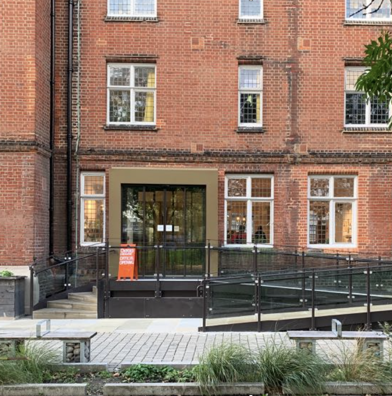 The outside of a red brick building with an access ramp and steps leading up to its black doors. A bright orange signboard stands at the entrance.