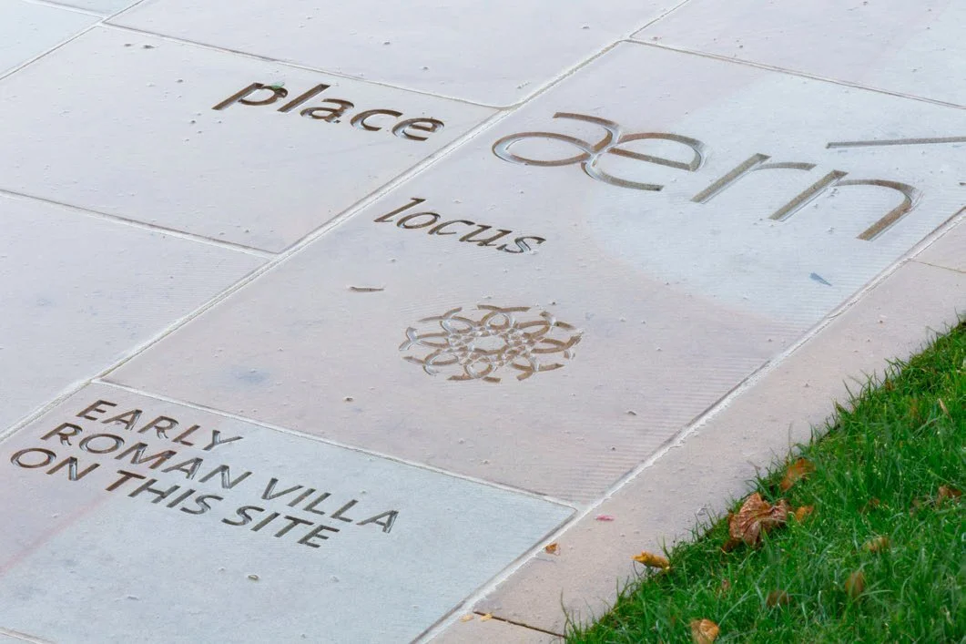 Close up of paving stones on a pathway with words and a circular symbol carved into them. The words are in different typefaces and read: “place”, “locus”, “aern”, and “Early Roman villa on this site.”