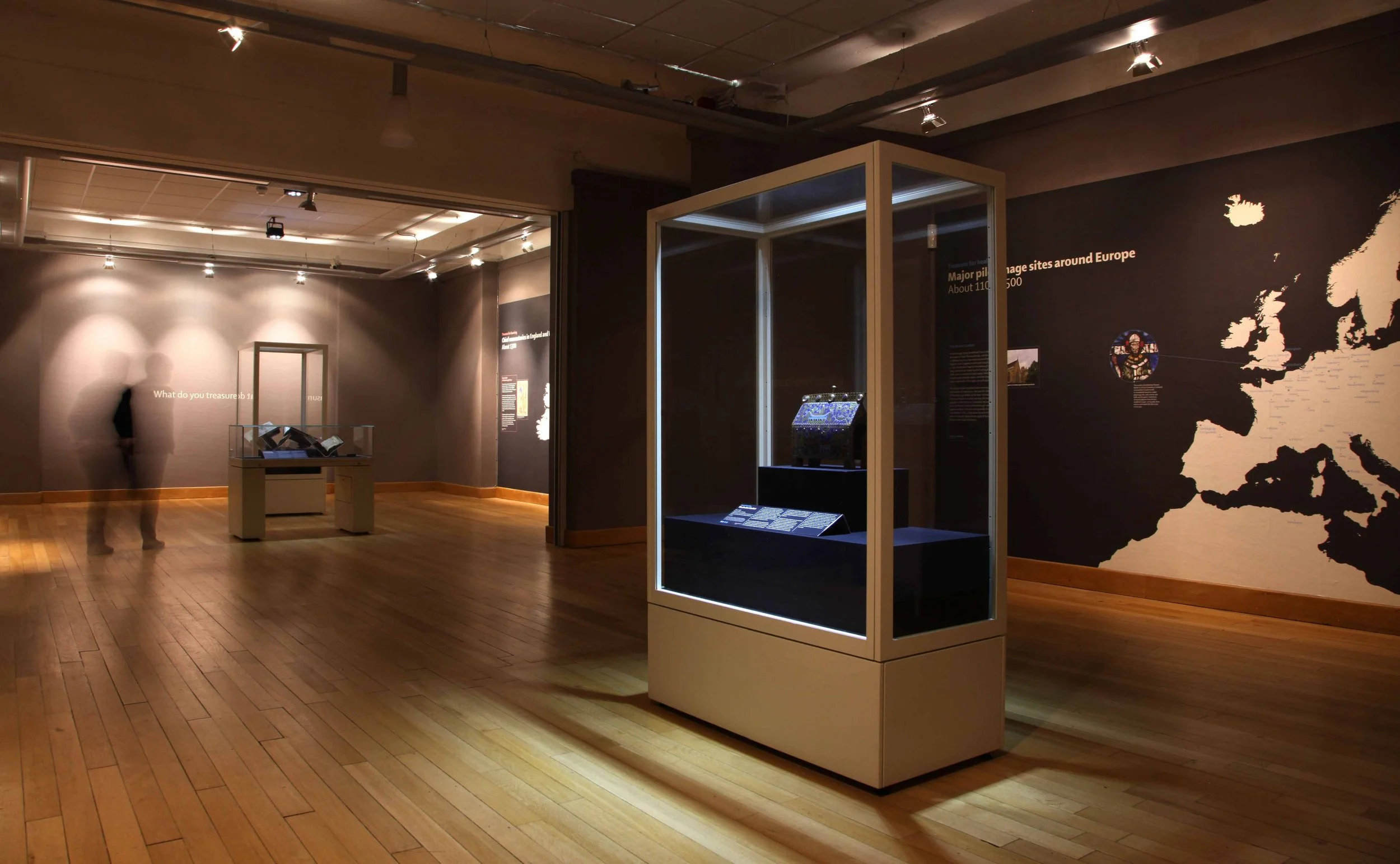 A visitor walks through a gallery with wooden floors and spotlights. Large minimalist black and white maps on the walls show medieval pilgrimage sites in Europe. Three showcases in the centre of the gallery display open books and objects.