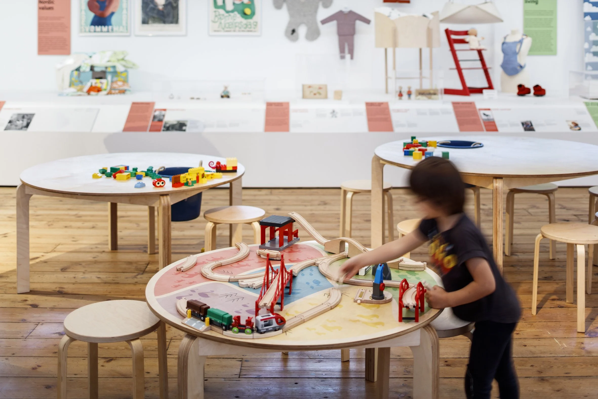 A child plays with a wooden train set at a round wooden table. Two similar tables behind it offer building bricks and Lego. In the background, framed posters, clothing and objects are displayed on a long white plinth with pink object labels.