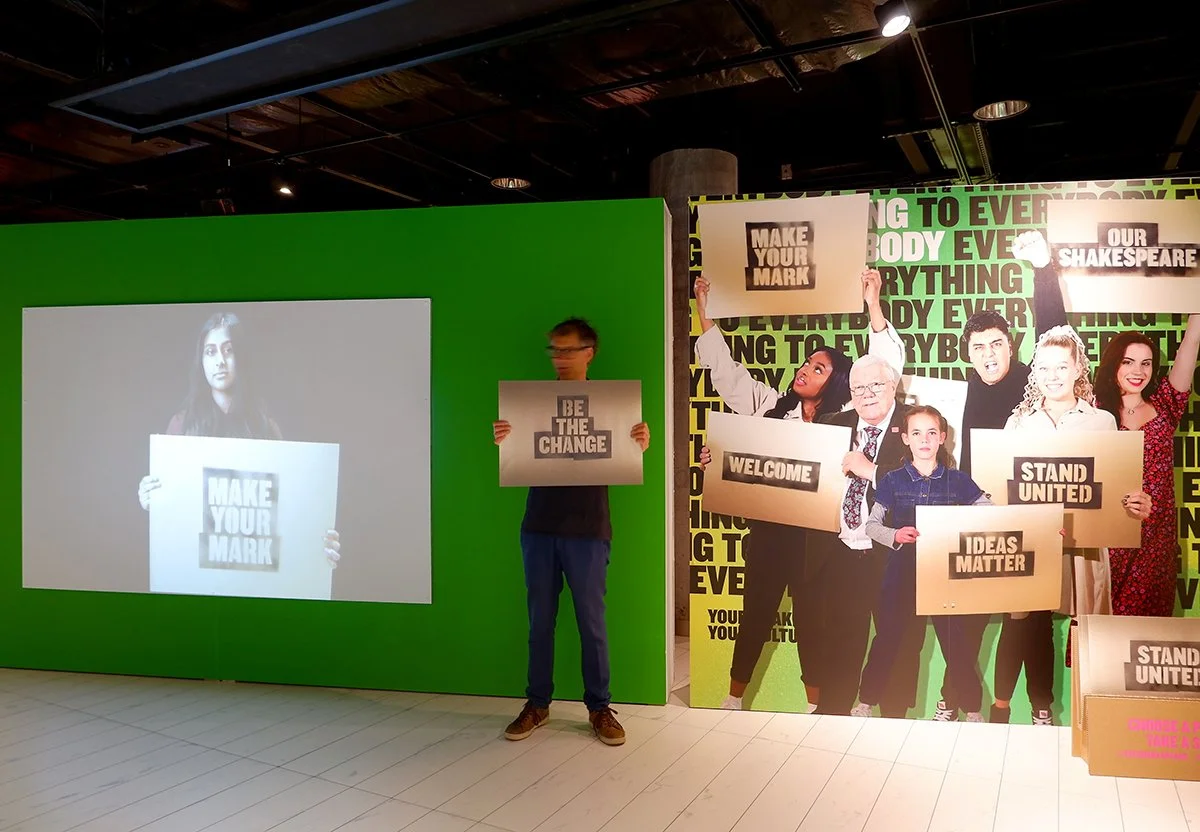 A man holds a protest placard in front of a video projection of a person holding a similar sign. To their right is a large image of a group of people also holding signs up.