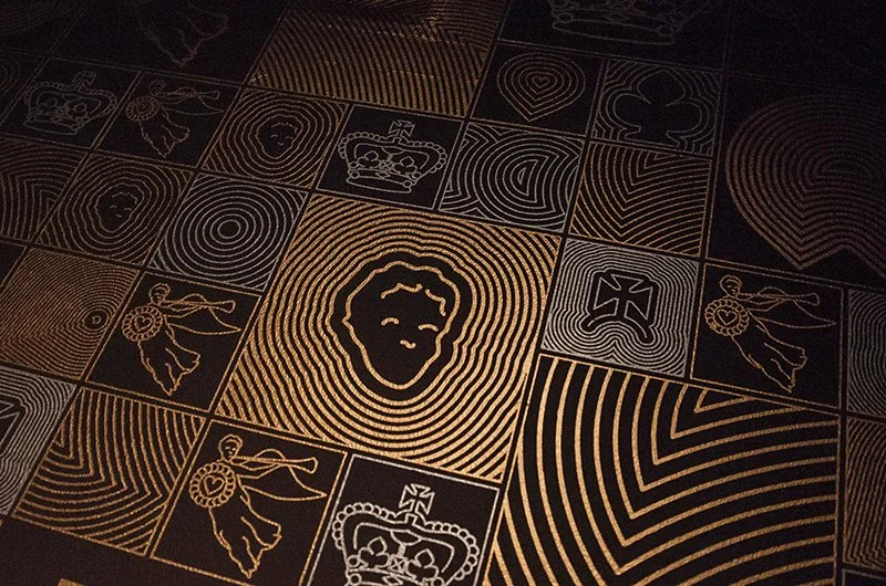 A close up of repeated motifs in squares of different sizes, including a figure with a trumpet, circular lines, a cross and a crown. The background is black and the motifs are printed in glittering silver and gold.