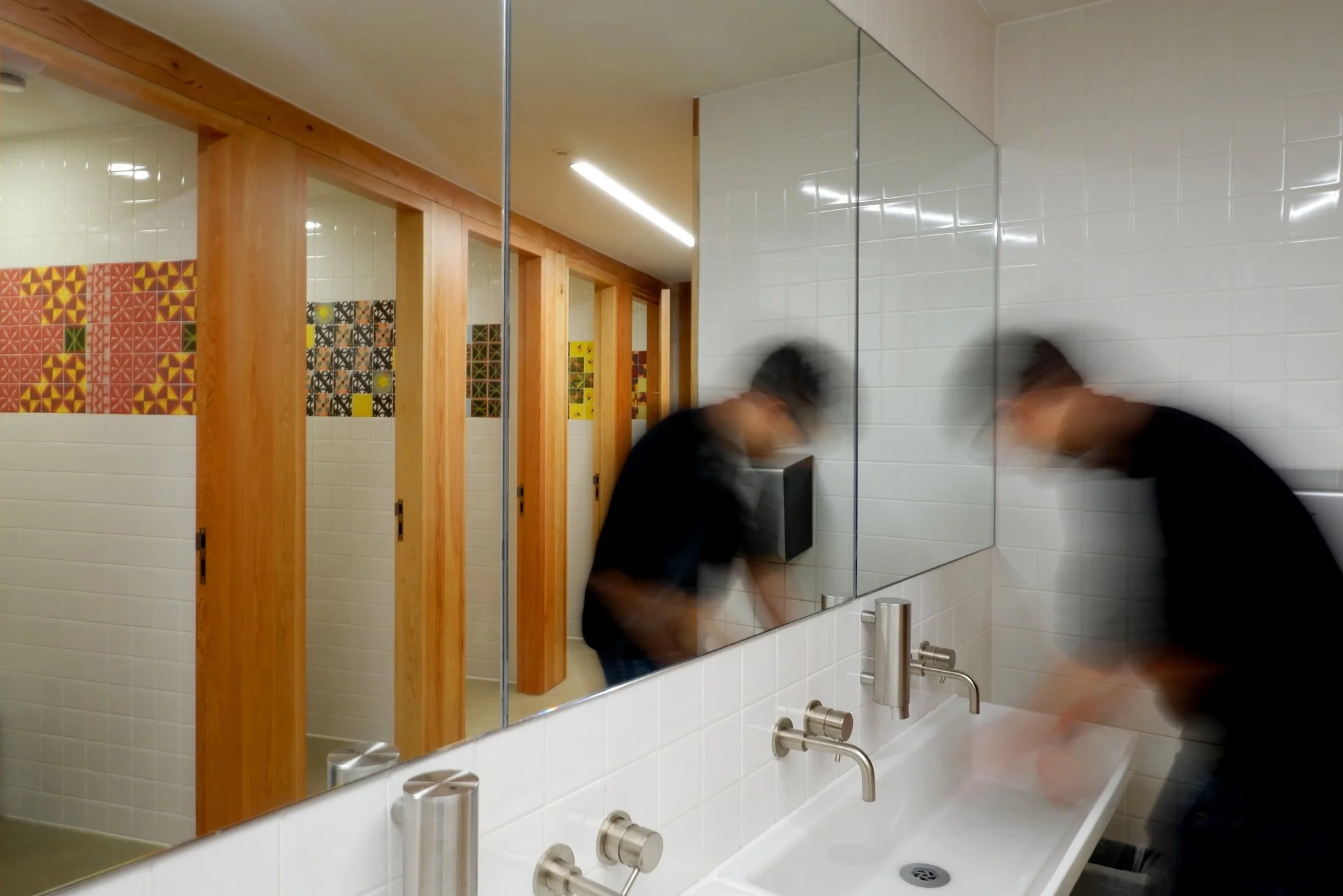 A man washes his hands in a white tiled bathroom. A large mirror in front of him reflects the toilet stalls behind him. Each has a colourful strip of patterned tiles on the walls.