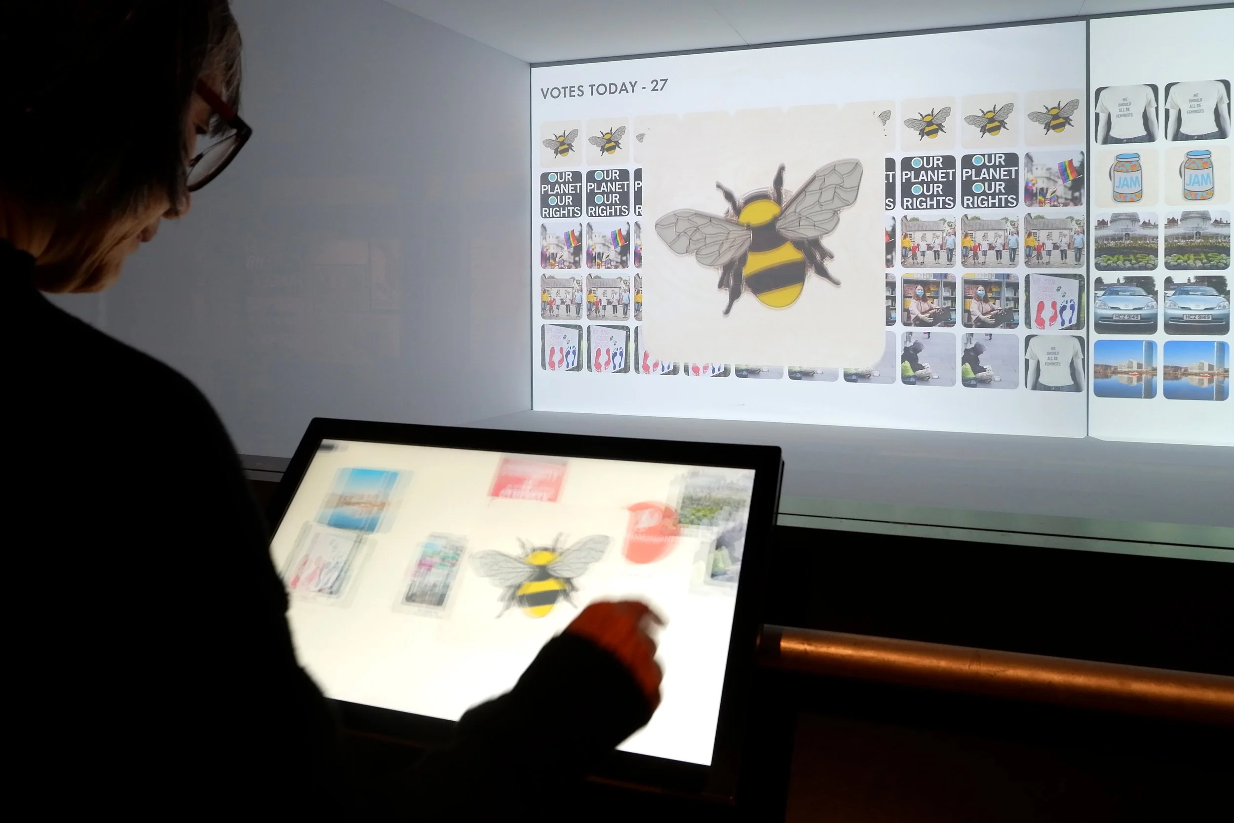 A visitor uses a touchscreen to select an image of a bumblebee. A projected image in front of them has the words "votes today" and shows the bee alongside other photos.