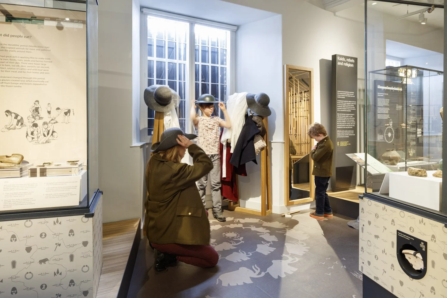 A family try on hats at a dressing up interactive station in a gallery surrounded by exhibition showcases. The floor is decorated with playful motifs of a mammoth.