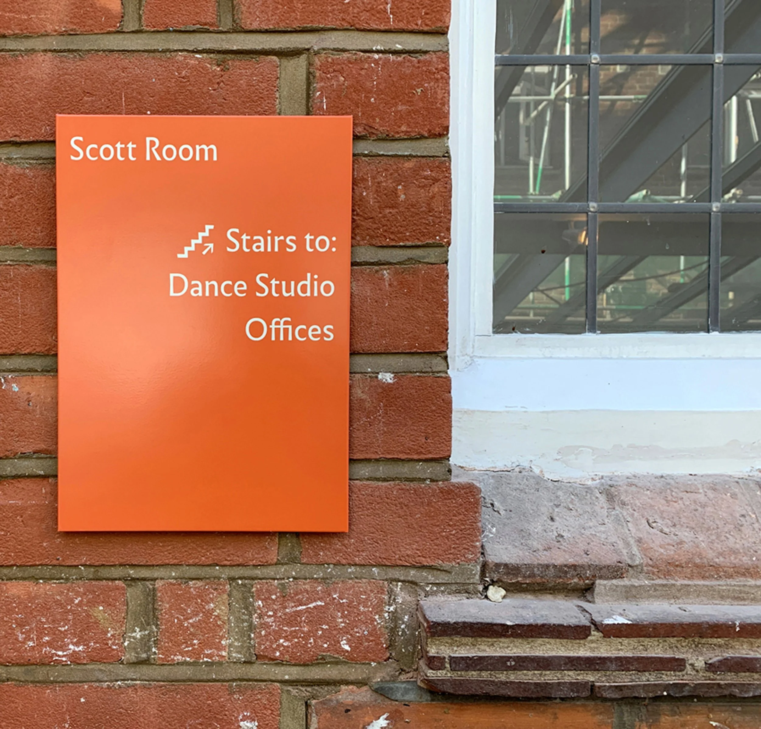 An orange wayfinding sign with white text mounted on a red-brick wall next to a window. The text reads: “Scott Room” with an icon of stairs leading up, and the words “Stairs to: Dance Studio. Offices.”